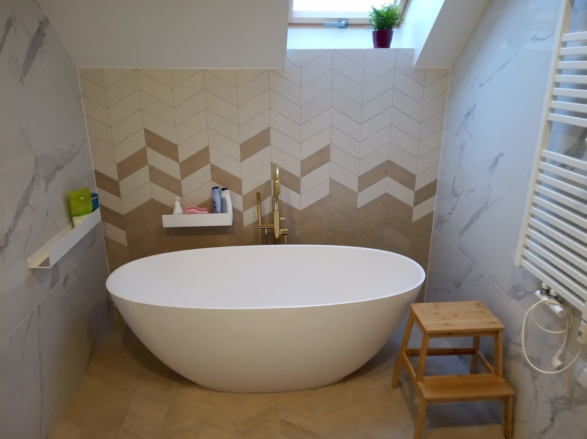 Nowoczesna łazienka z wolnostojącą wanną, mozaiką w jodełkę na ścianie i drewnianym stołkiem. Minimalistyczny design, jasne kolory i naturalne światło.