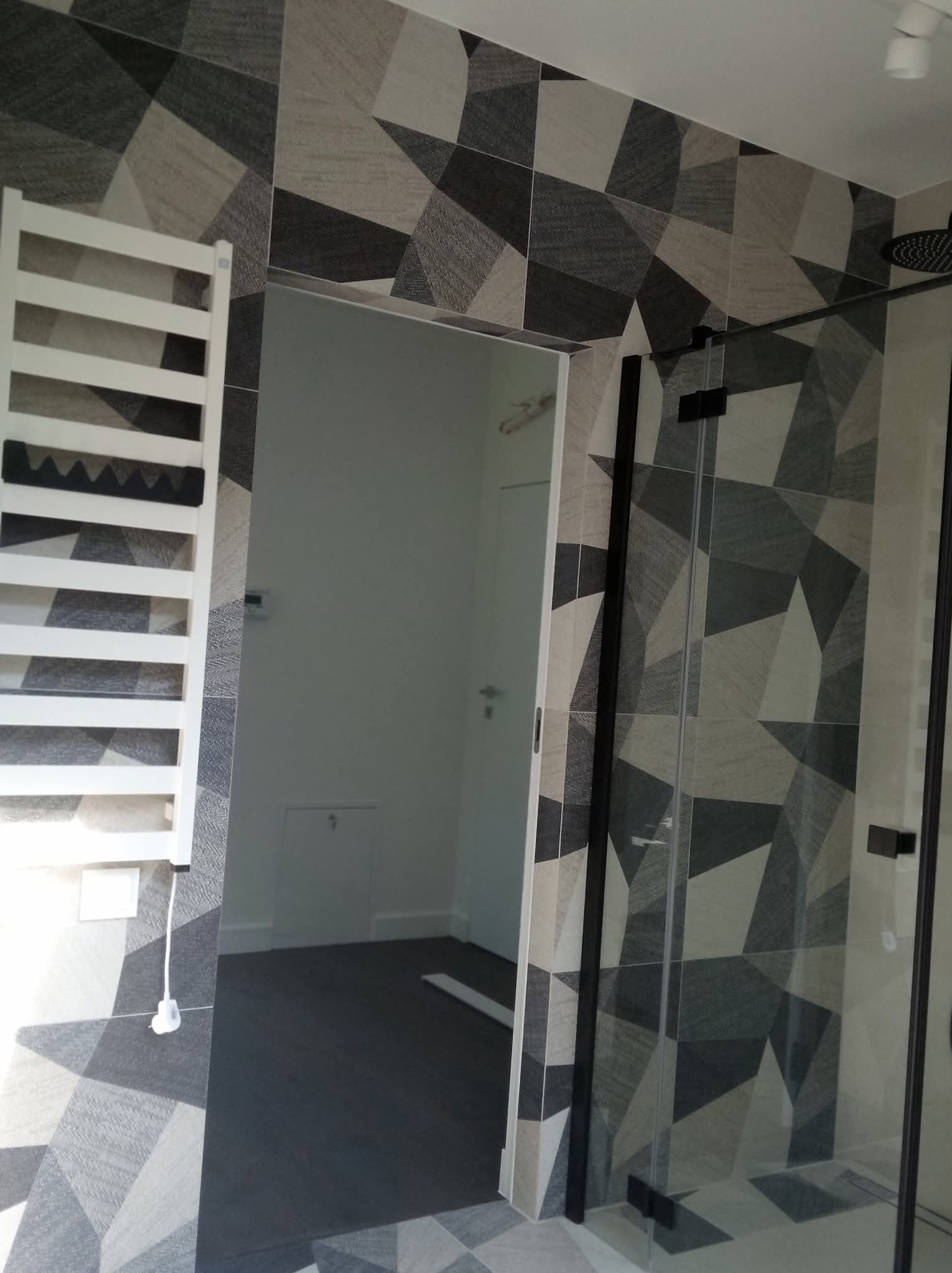 Wykończona łazienka z geometrycznymi płytkami w odcieniach szarości i bieli, kabina prysznicowa z czarnymi profilami, grzejnik drabinkowy. Nowoczesny design.
