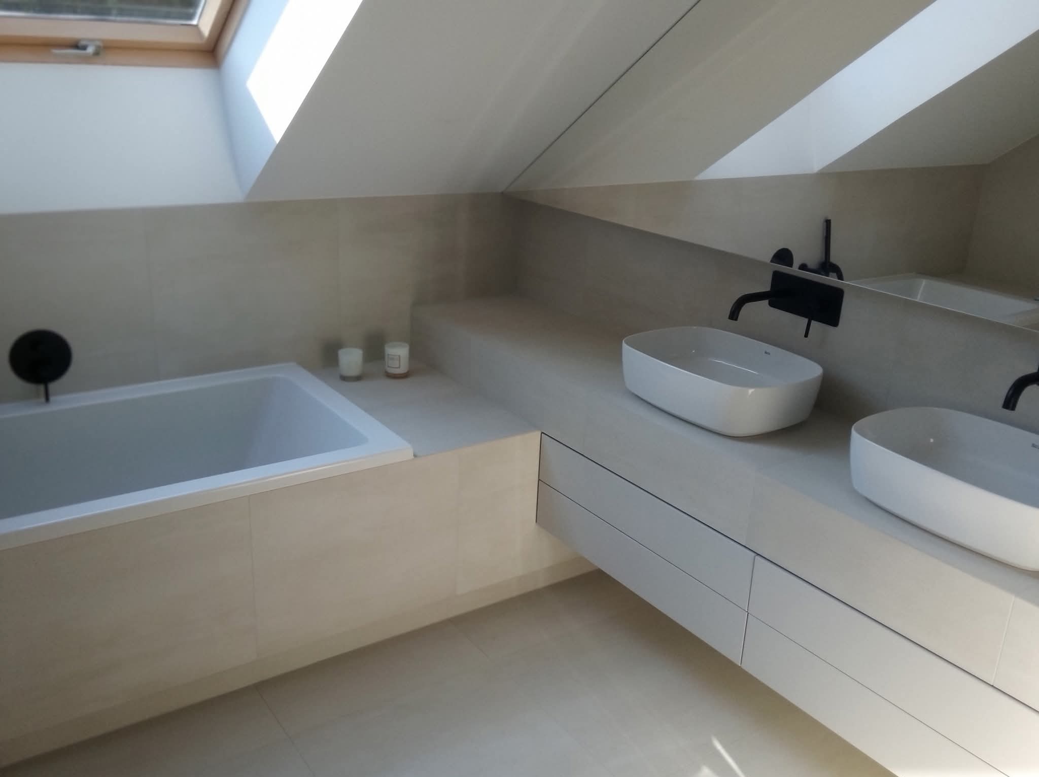 Jasna łazienka na poddaszu ze skośnym sufitem i oknem dachowym. Wanna obudowana płytkami, dwie umywalki nablatowe z czarną armaturą. Minimalistyczny design.