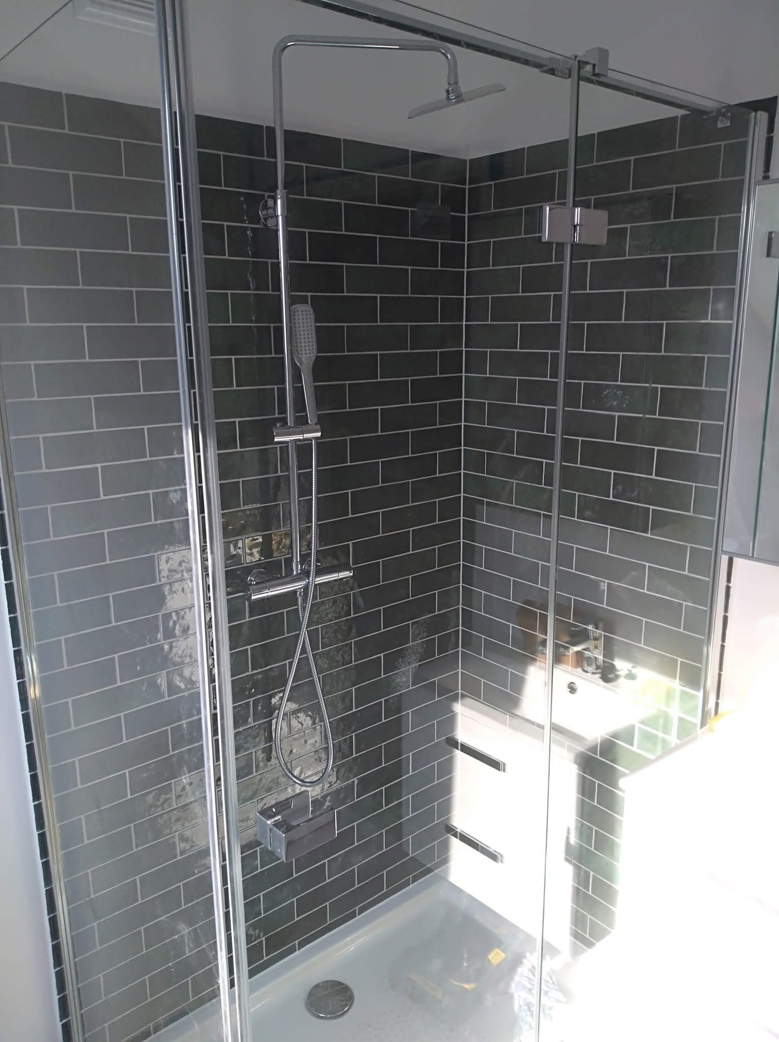 Nowoczesna kabina prysznicowa z szarymi, błyszczącymi płytkami ułożonymi w cegiełkę, chromowaną armaturą i szklanymi drzwiami. Widoczny odpływ liniowy i fragment umywalki.