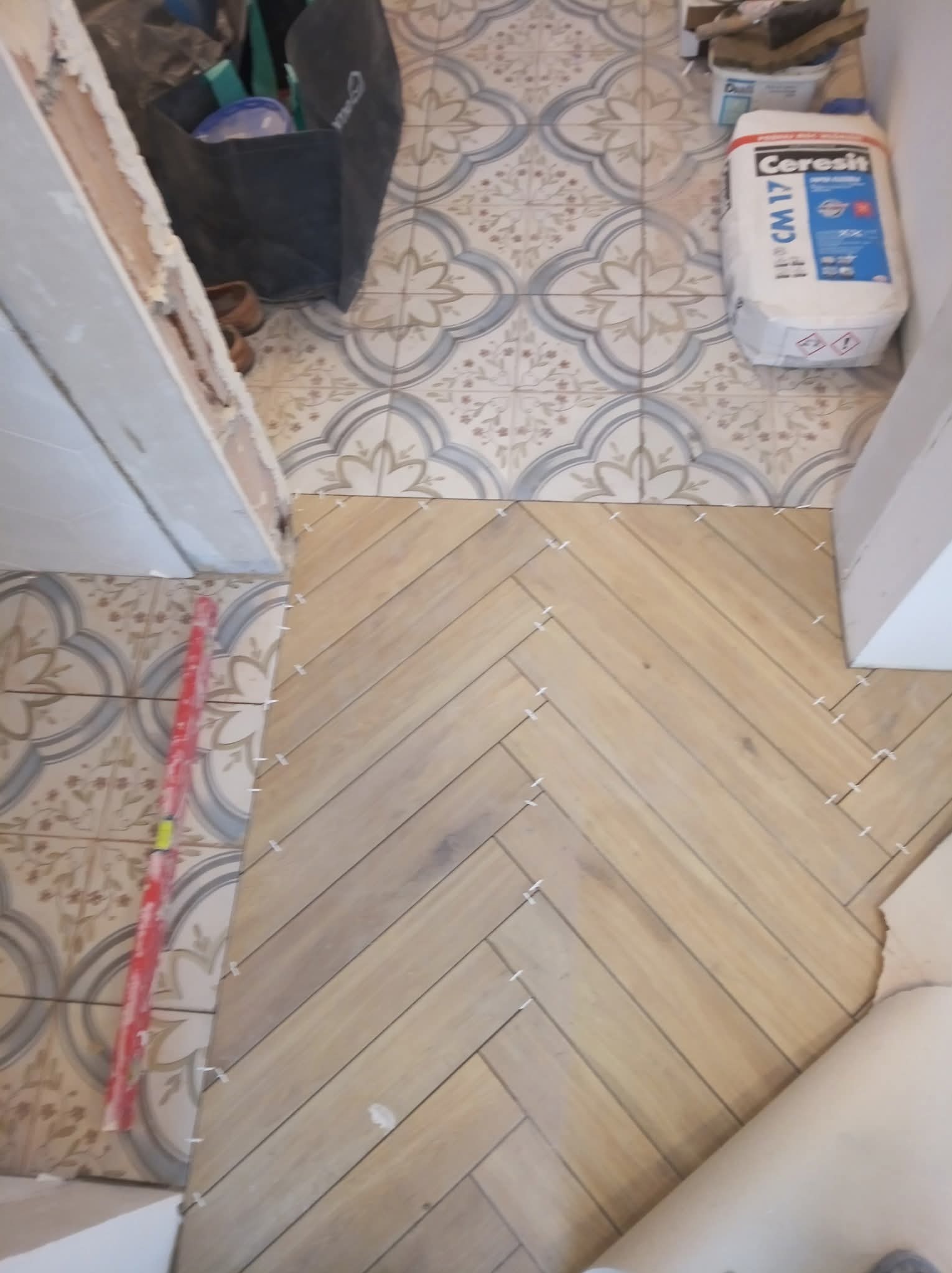 Układanie parkietu w jodełkę, widoczne płytki z krzyżykami dystansowymi, częściowo ułożone na tle wzorzystej podłogi. Paczka kleju do płytek w tle.