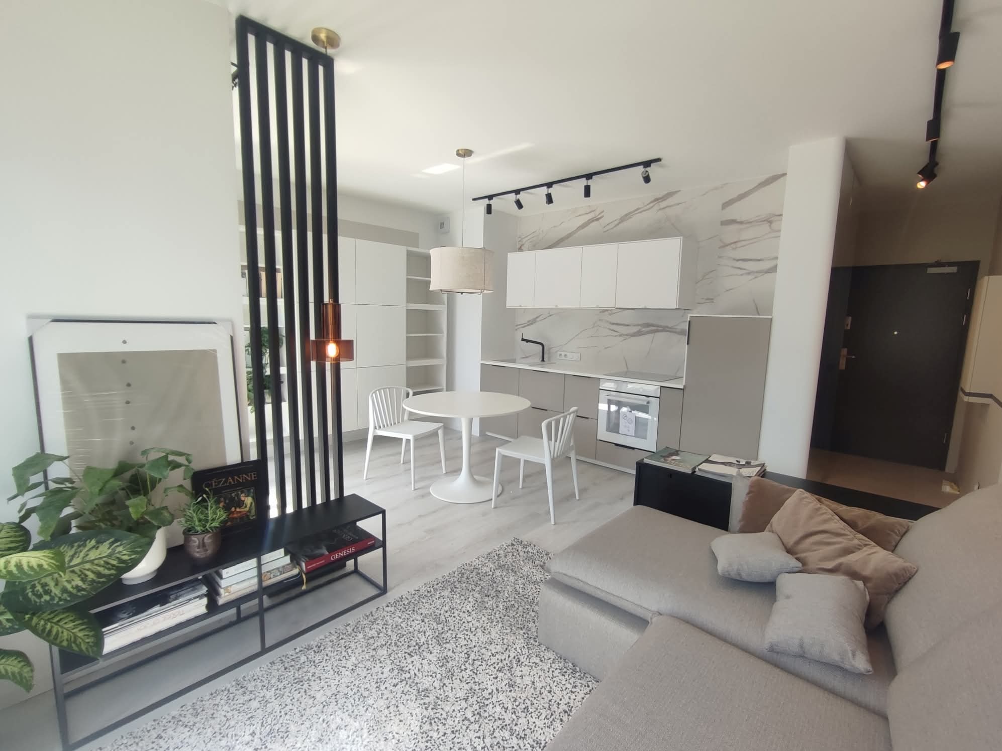 Jasne wnętrze salonu z aneksem kuchennym: marmurowa ściana, białe meble, okrągły stół i szara sofa. Elementy dekoracyjne: rośliny, książki, lampy.