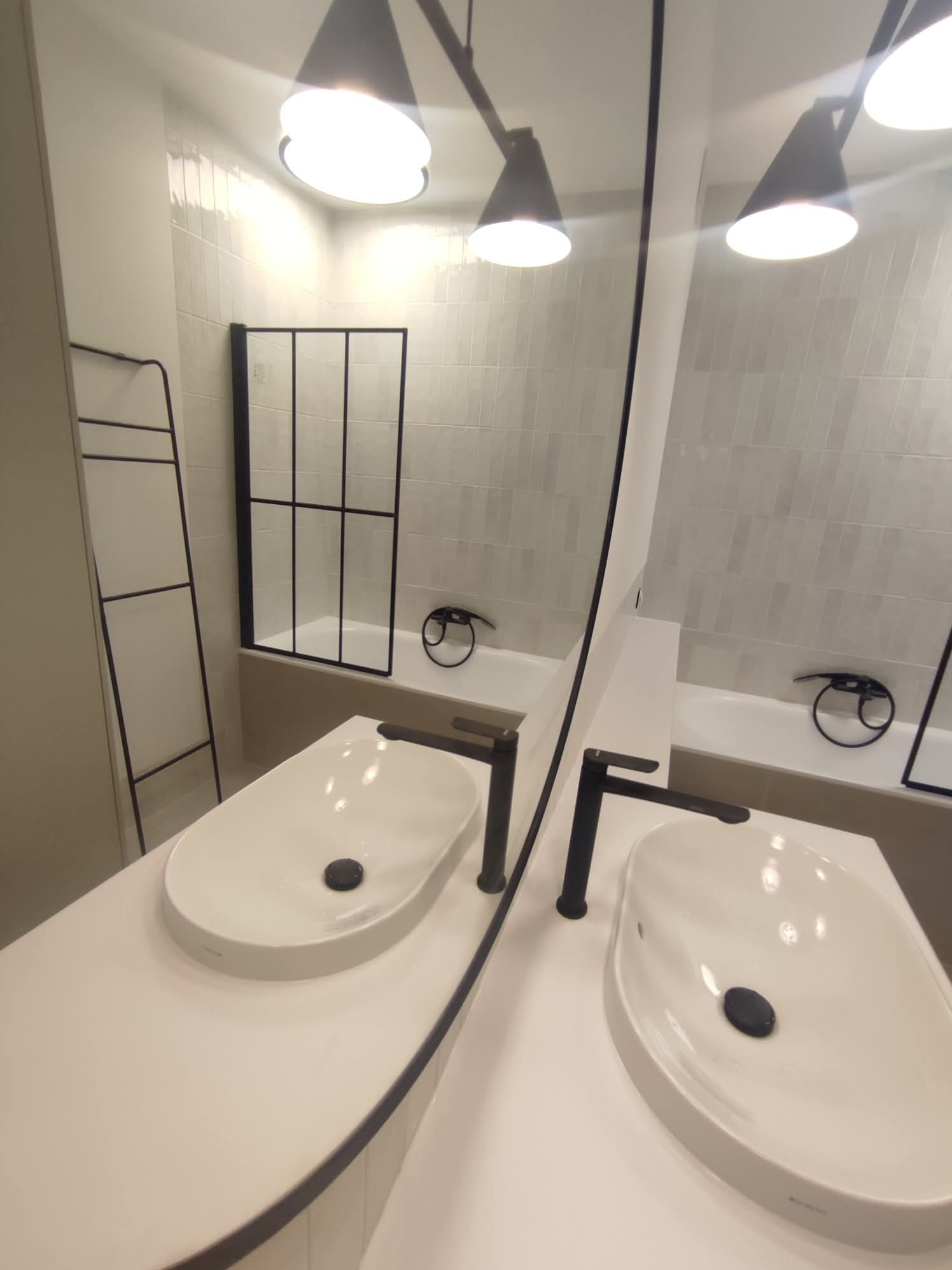 Nowoczesna łazienka z owalną umywalką nablatową, czarną armaturą i kabiną prysznicową w tle. Minimalistyczny design, jasne płytki i oświetlenie punktowe.