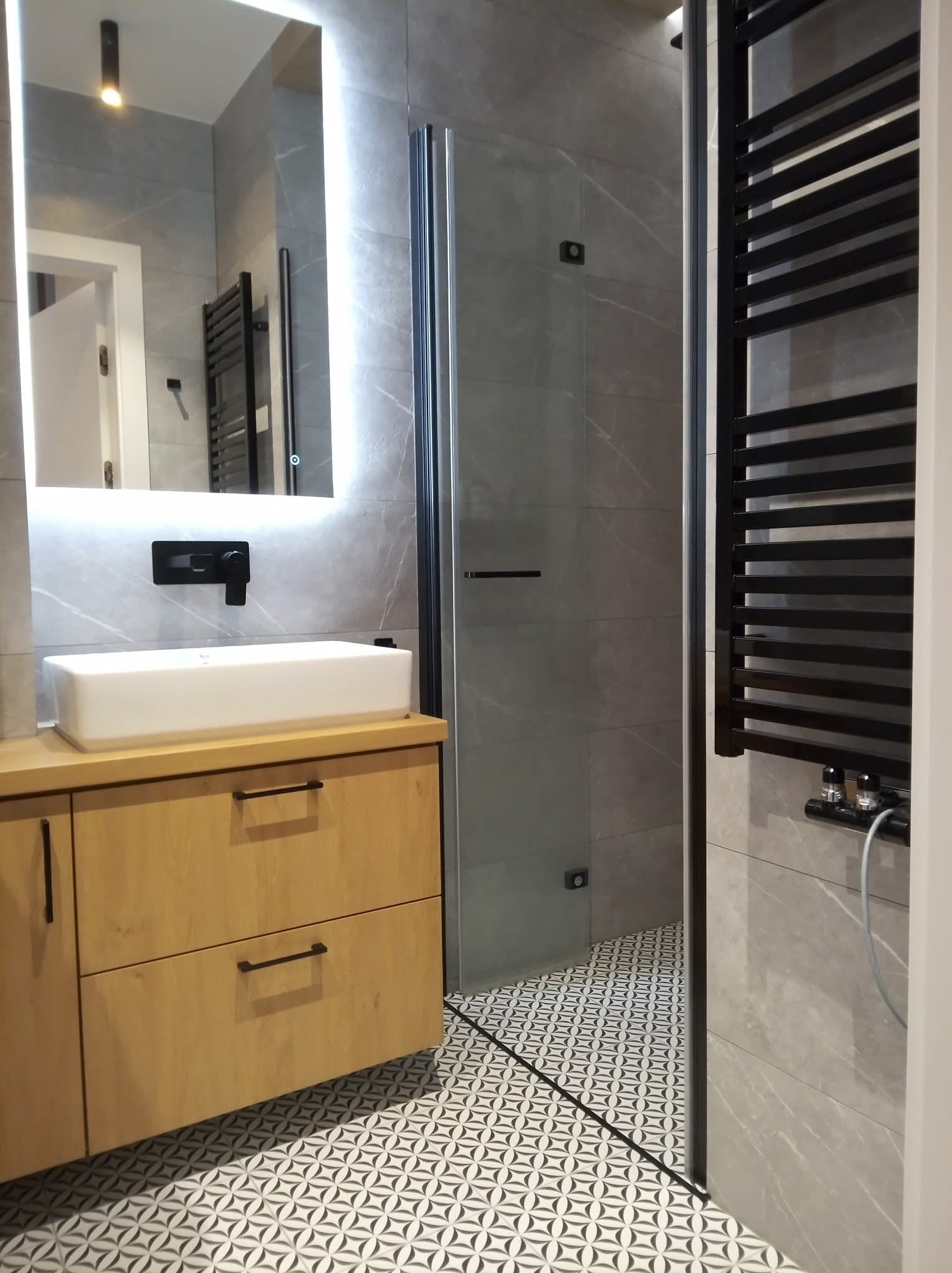 Nowoczesna łazienka z drewnianą szafką pod umywalką, szarą glazurą imitującą marmur i kabiną prysznicową. Czarny grzejnik drabinkowy i wzorzysta podłoga.