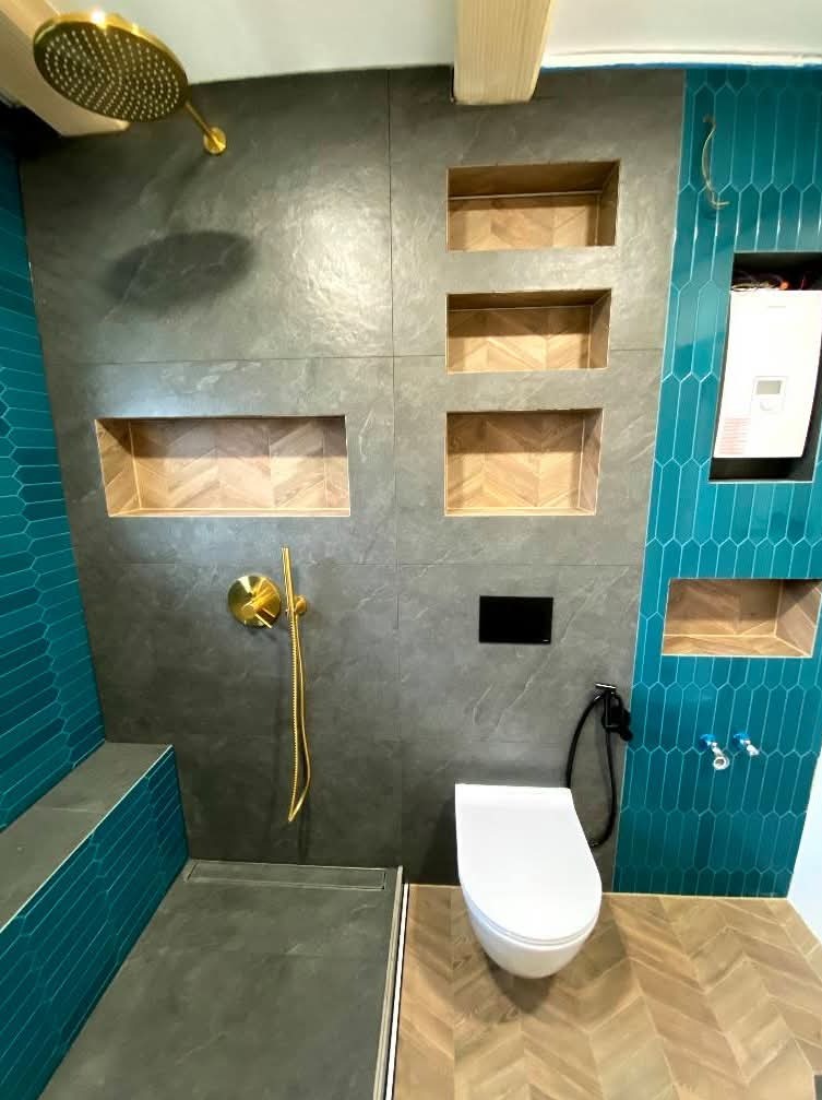 Nowoczesna łazienka po remoncie: szare płytki, złota armatura, turkusowe akcenty i wnęki z drewnianym wzorem jodełki. Uwagę przykuwa deszczownica i minimalistyczna toaleta.