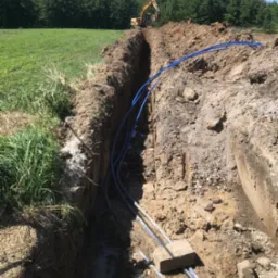 Długi, wąski wykop w ziemi z ułożonymi niebieskimi rurami, w tle widoczny żółty koparka pracująca na końcu wykopu, po bokach trawa i drzewa.