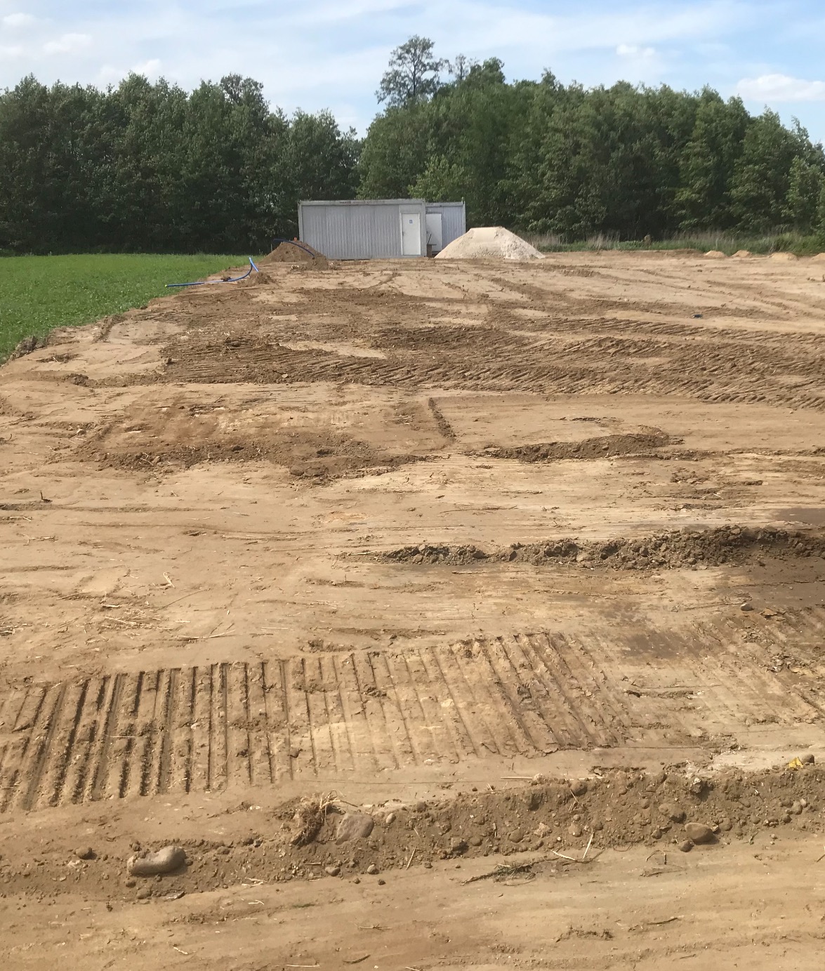 Przygotowany teren pod budowę z widocznymi śladami maszyn, kontener biurowy w tle i hałda piasku.