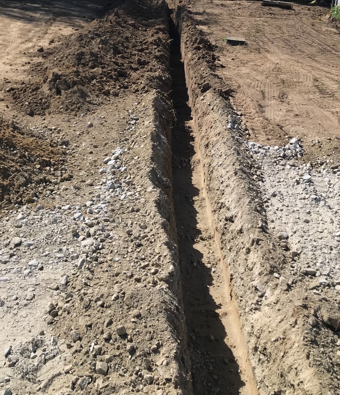 Wąski rów wykopany w ziemi, widoczne ślady opon w tle, ziemia usypana po obu stronach wykopu.