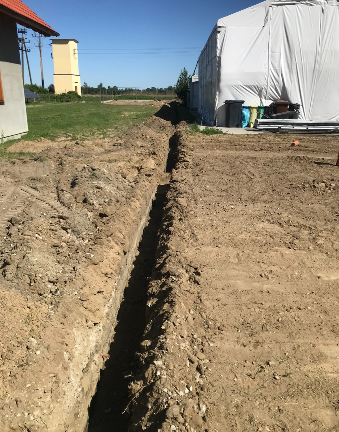Długi, wąski rów wykopany w ziemi, widoczny na tle budynku gospodarczego i transformatora, z rozrzuconą ziemią po bokach.