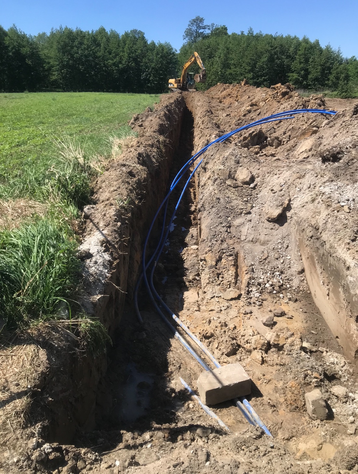 Żółta koparka w tle wykopu pod instalacje z ułożonymi niebieskimi rurami, zabezpieczonymi betonowym blokiem.