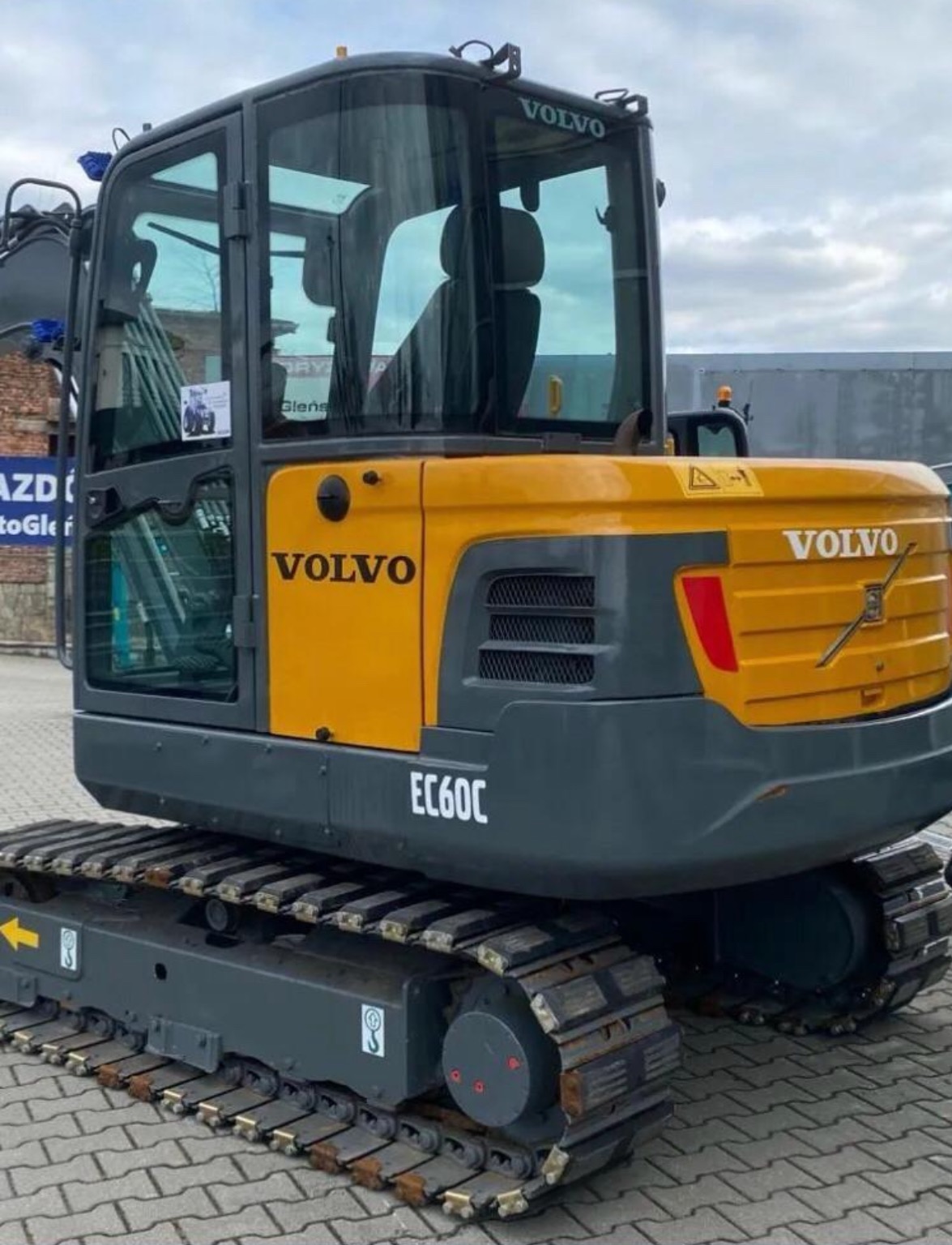 Żółto-szara minikoparka Volvo EC60C z kabiną, stojąca na kostce brukowej przed budynkiem z reklamą.
