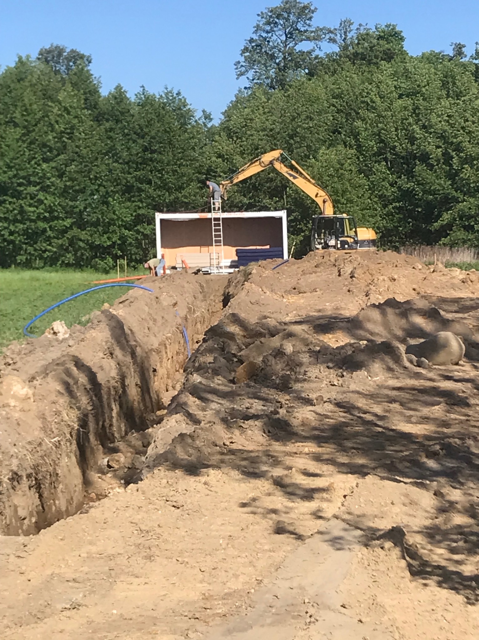 Wykop w Prusicach z ułożonym niebieskim przewodem, w tle kontener budowlany i koparka, dwóch pracowników przy kontenerze.