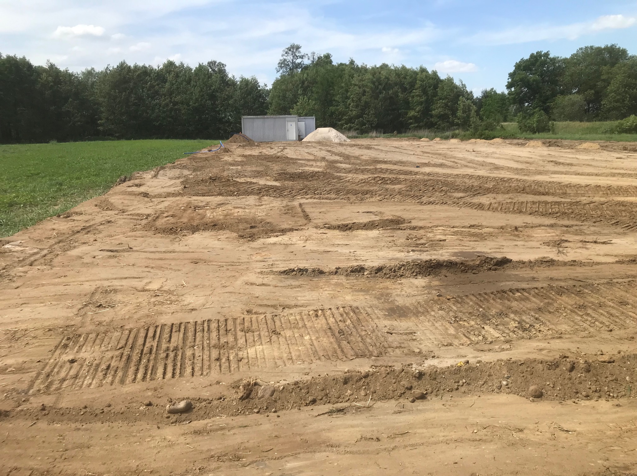 Przygotowany teren pod budowę z widocznymi śladami maszyn, w tle kontener budowlany i pryzma piasku, otoczony zielenią.