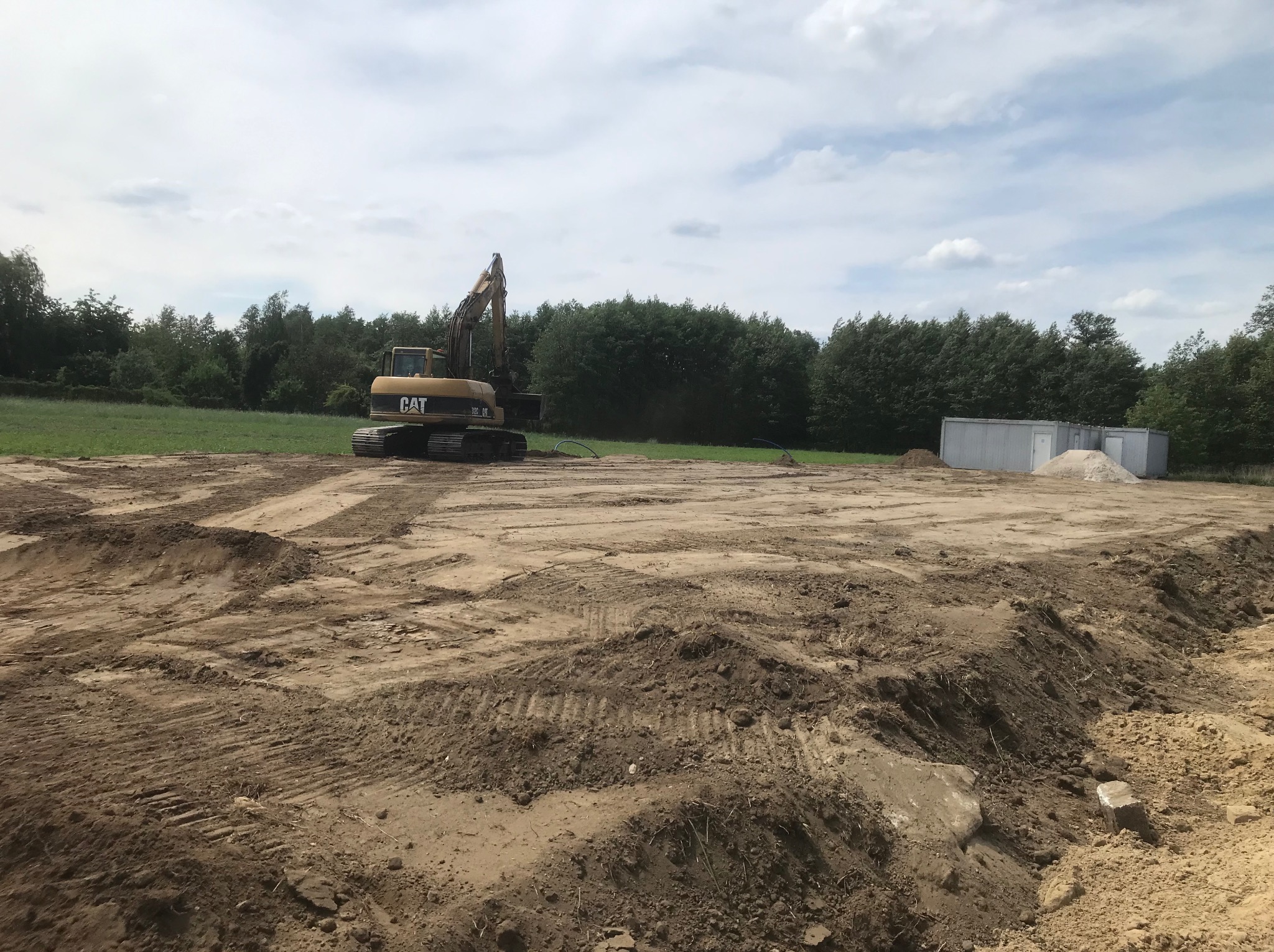 Żółta koparka CAT na przygotowanym placu budowy z widocznymi śladami opon i wyrównanym terenem, w tle zieleń drzew i kontener biurowy.