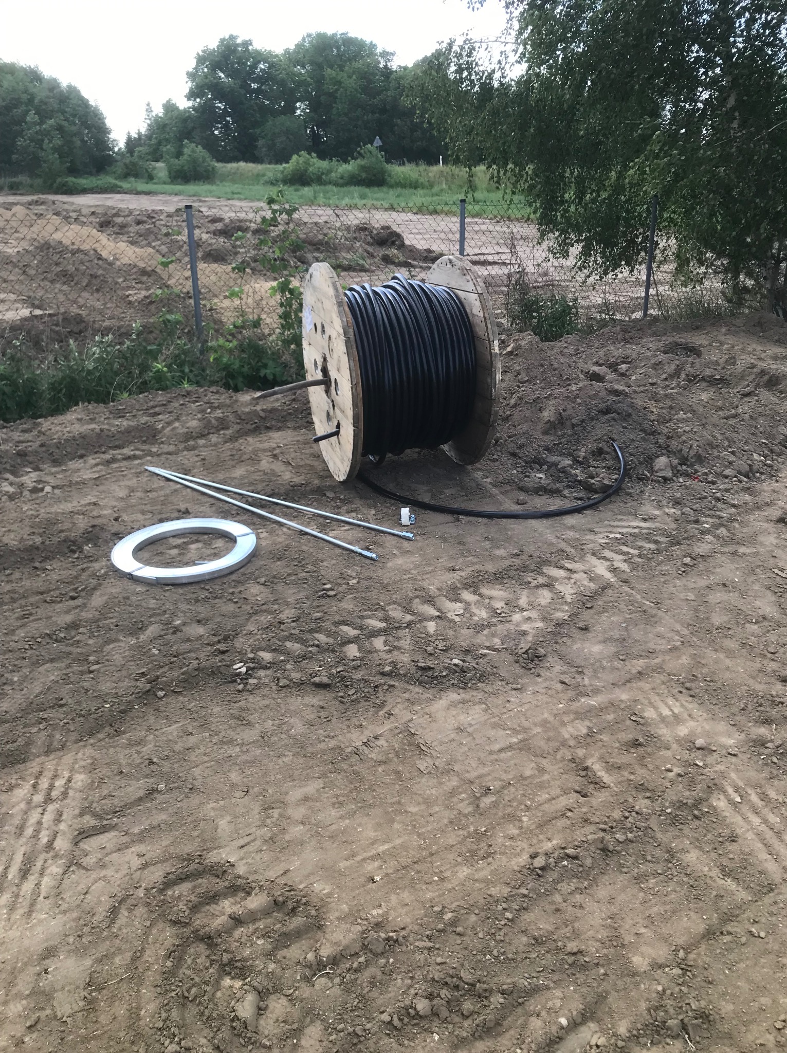 Szeroka szpula z nawiniętym czarnym kablem leżąca na ziemi obok metalowych prętów i okrągłej obejmy, w tle wykopany teren z siatką ogrodzeniową.