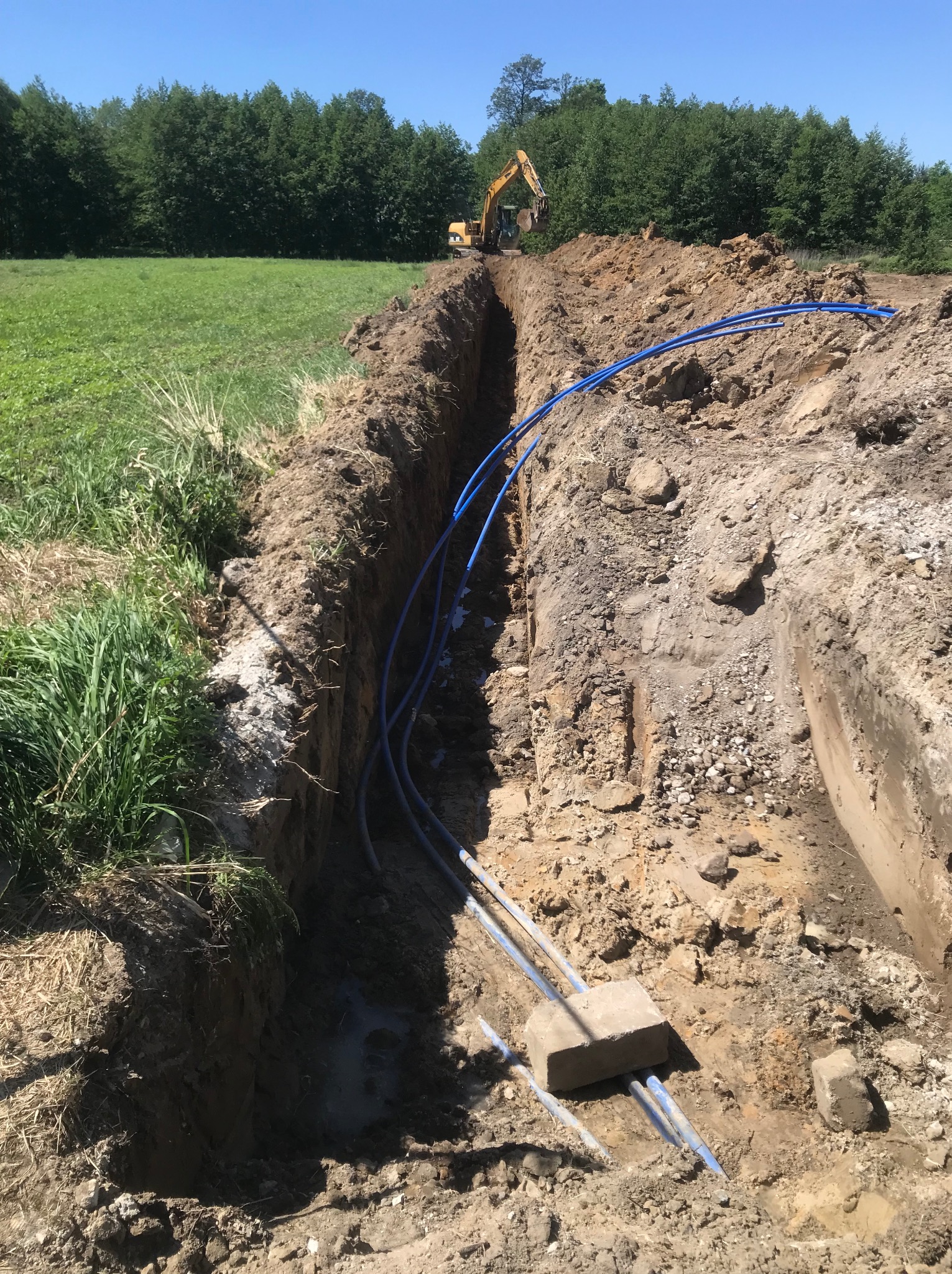Długi, wąski wykop w ziemi z ułożonymi niebieskimi rurami, w tle widoczny żółty koparka pracująca na końcu wykopu, po bokach trawa i drzewa.