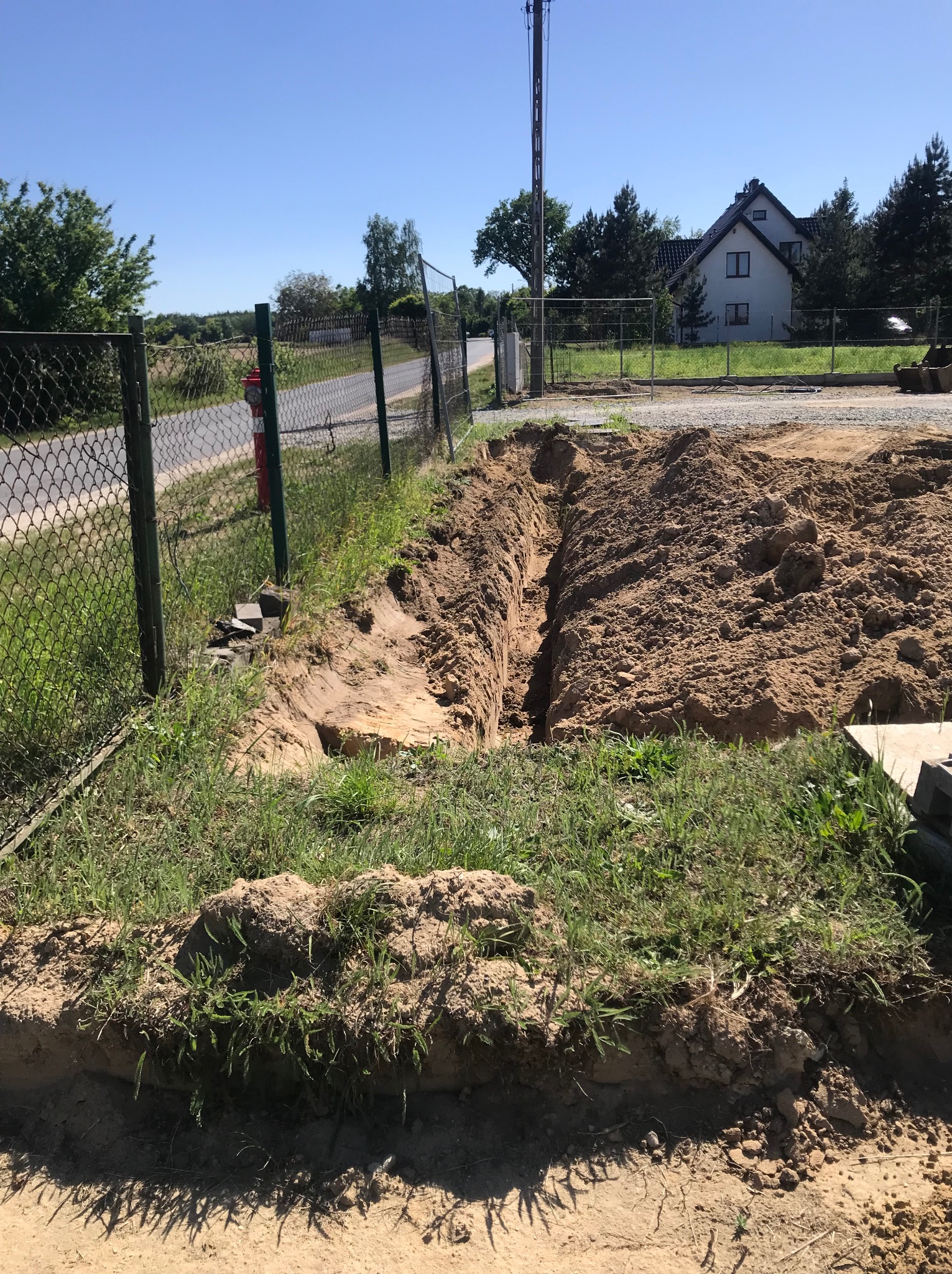 Wykopany wąski rów w ziemi wzdłuż ogrodzenia z siatki, z hałdą piasku obok, teren budowy z domem w tle.