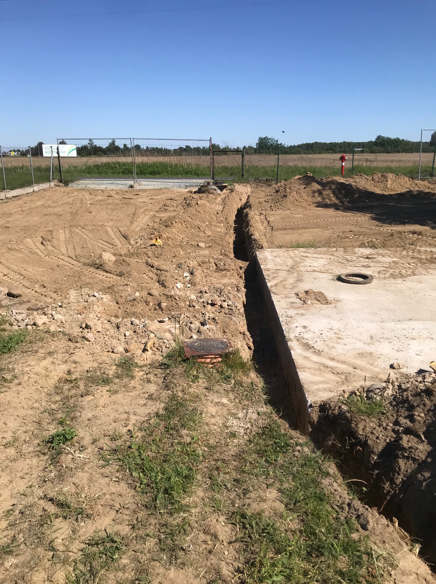 Wykonany wykop pod fundamenty z widocznym betonowym elementem konstrukcyjnym i studzienką kanalizacyjną na terenie budowy, w tle widoczne ogrodzenie i krajobraz.