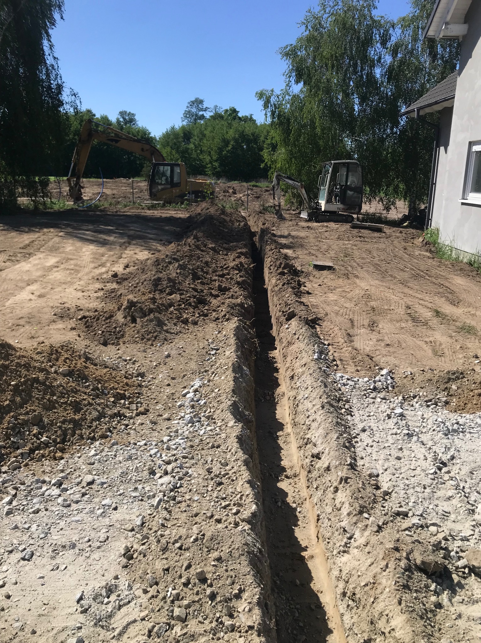 Długi, wąski wykop w ziemi z dwoma koparkami w tle, przygotowany pod instalację, widok z perspektywy ziemi, jasne światło słoneczne.