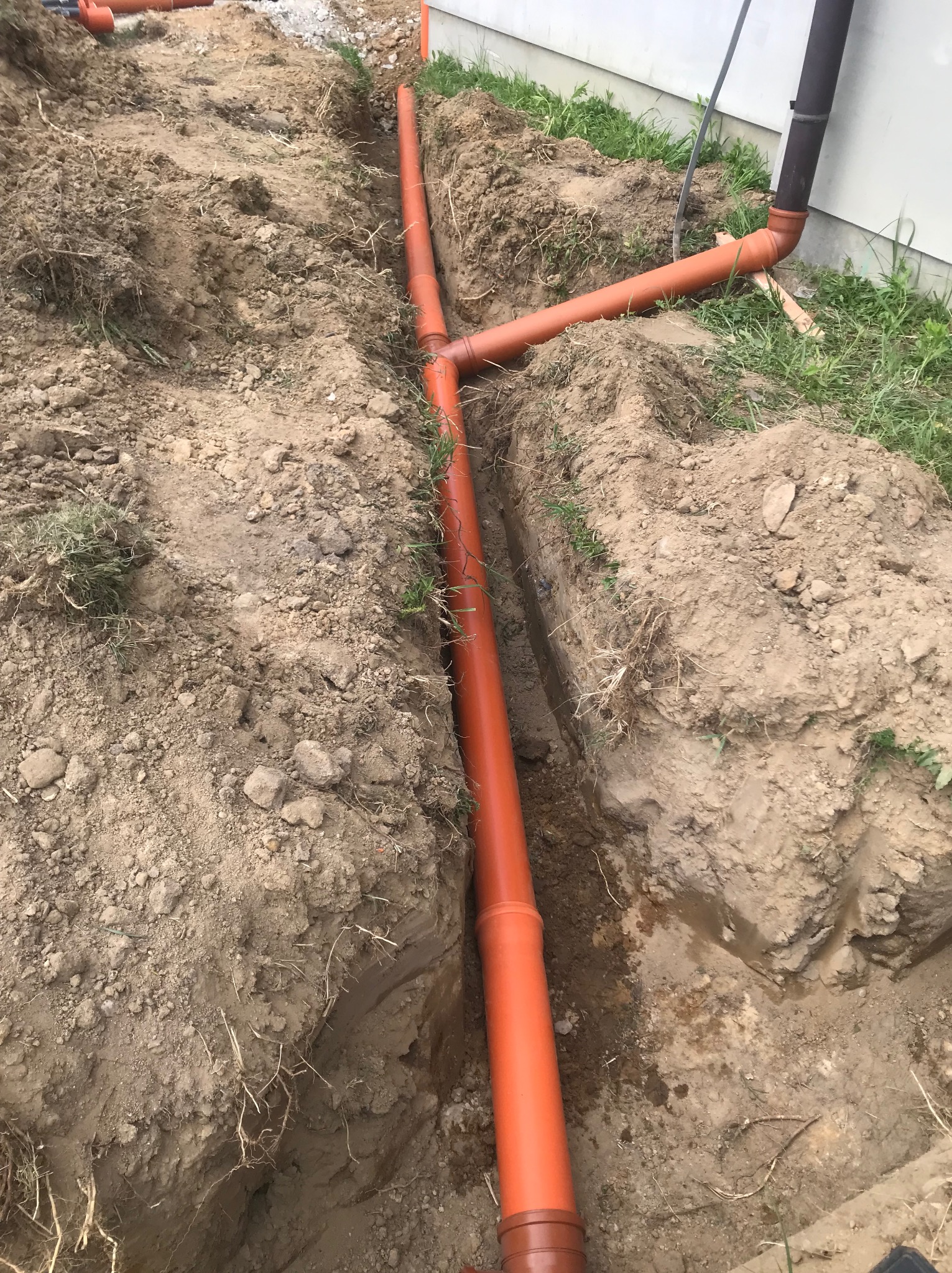 Pomarańczowe rury kanalizacyjne ułożone w wykopie ziemnym wzdłuż ściany budynku, widoczne złącza i odgałęzienia, ziemia lekko wilgotna.