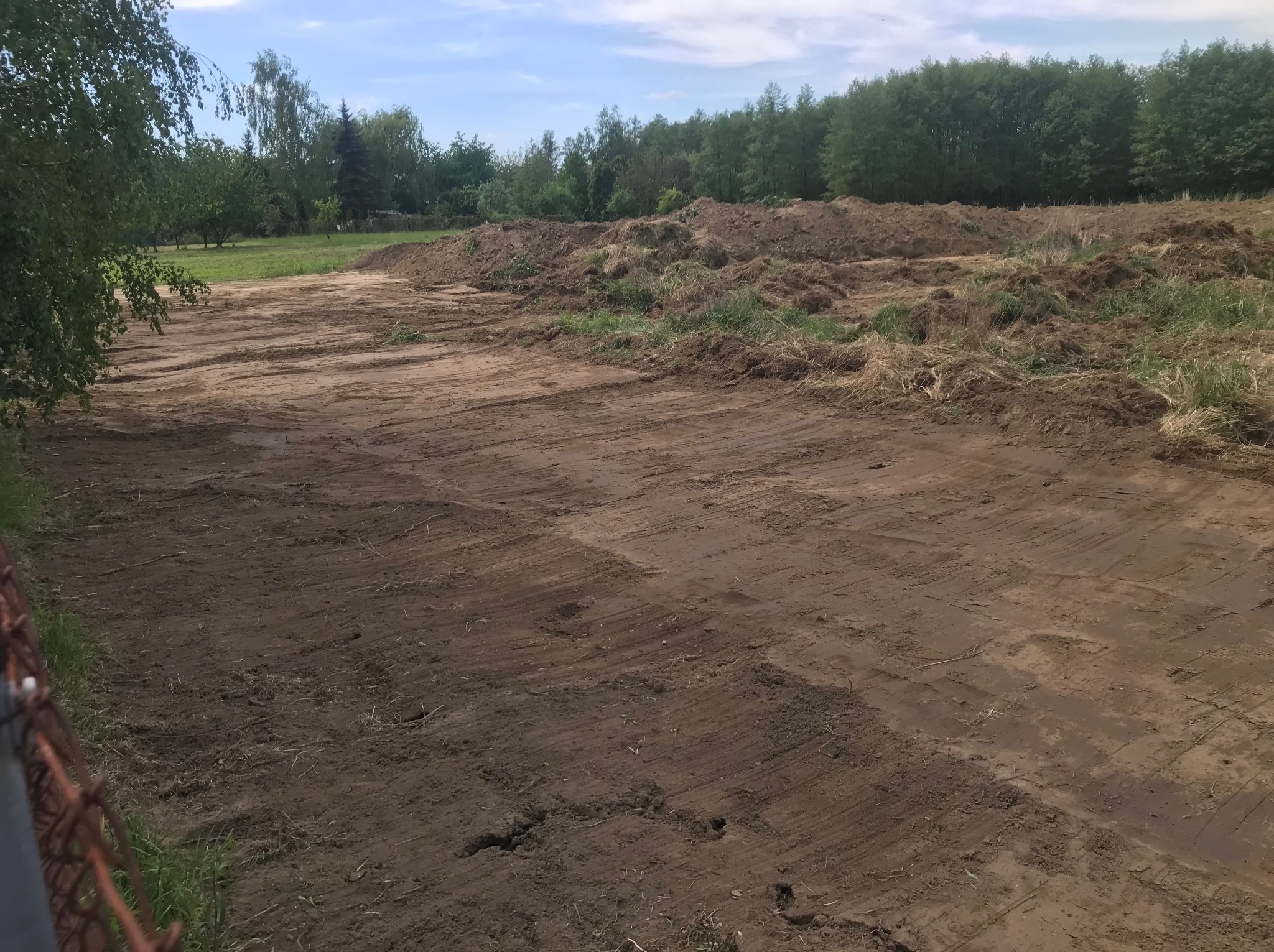 Przygotowany teren pod budowę z widocznymi śladami po maszynach i pryzmami ziemi w tle, otoczony zielenią.