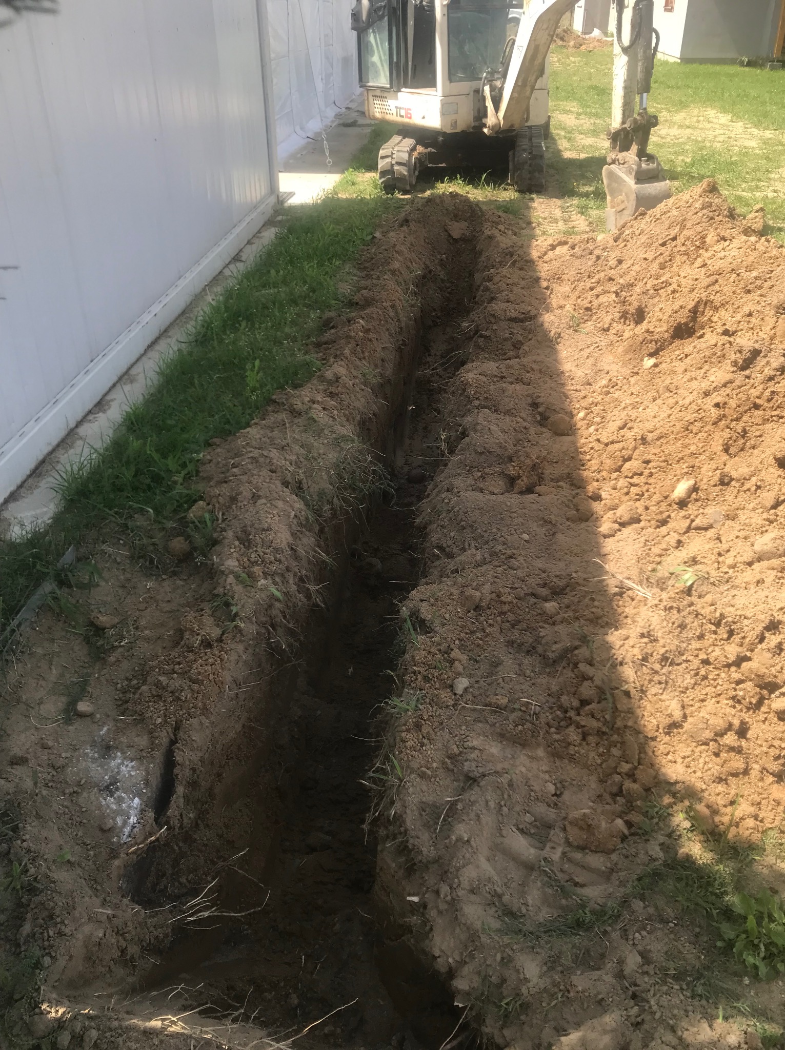 Długi, wąski wykop w ziemi, wykonany wzdłuż białego panelowego ogrodzenia, z małą koparką w tle i stertą ziemi po prawej stronie.