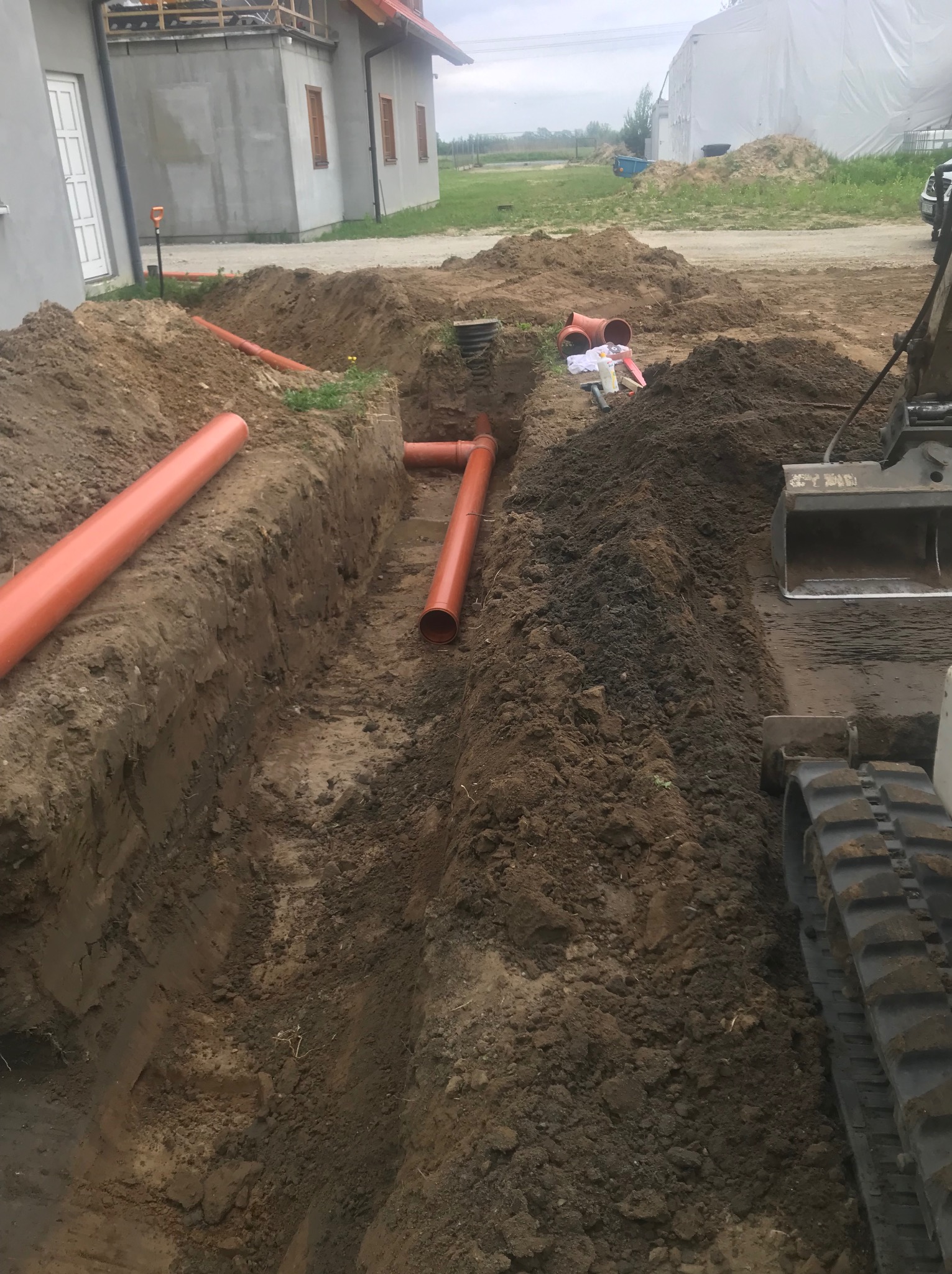 Wykopany rów z ułożonymi pomarańczowymi rurami kanalizacyjnymi, widoczna koparka i budynki w tle.