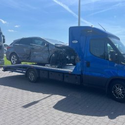 Pomoc drogowa Laweta 24h-OnlyCars - Niebieska ciężarówka z platformą transportową przewozi poważnie uszkodzony, szary samochód osobowy; widoczne zniszczenia karoserii i brak przedniej części pojazdu.