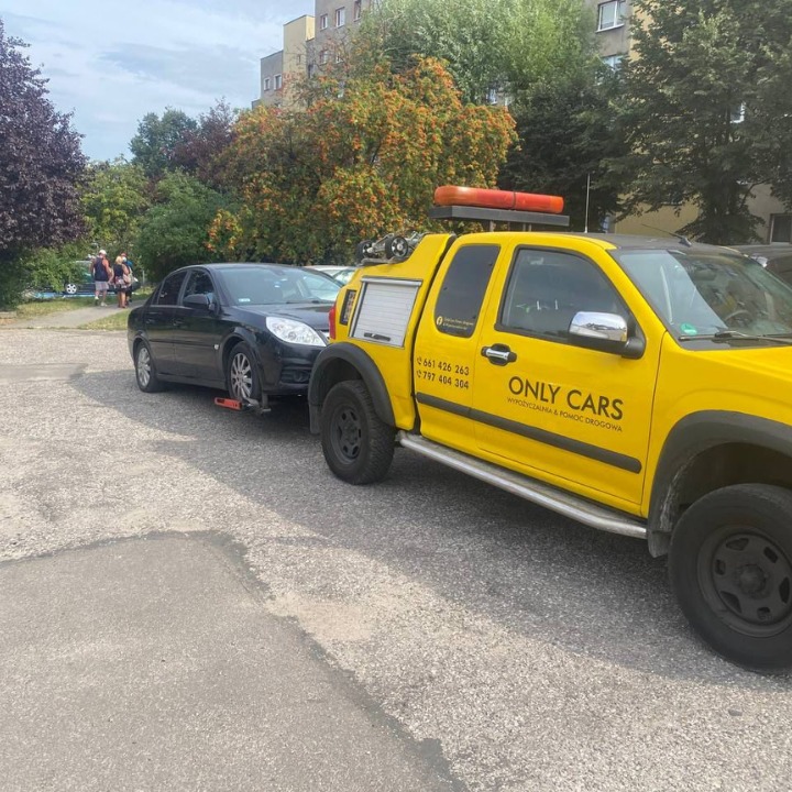 Żółta laweta firmy ONLY CARS z holowanym czarnym samochodem osobowym na parkingu przed budynkiem mieszkalnym.