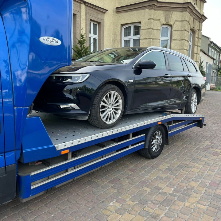 Czarny samochód kombi marki Opel Insignia na niebieskiej platformie lawety, widoczne uszkodzenia karoserii po stronie kierowcy.