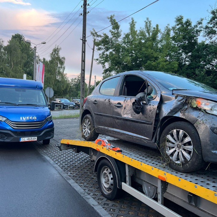 Uszkodzony, szary samochód osobowy Peugeot 207 z rozbitą szybą i wgnieceniami na karoserii, transportowany na żółtej lawecie. Widoczny niebieski samochód dostawczy Iveco z przodu lawety...