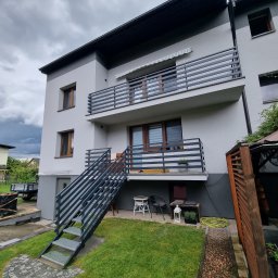 AR-Gelender Radosław Mojżyszek - Nowoczesny, dwukondygnacyjny dom z szarą elewacją, balkonami z metalowymi balustradami i zewnętrznymi schodami z metalu prowadzącymi na wyższy poziom. Widoczny fragment ogrodu z trawnikiem...