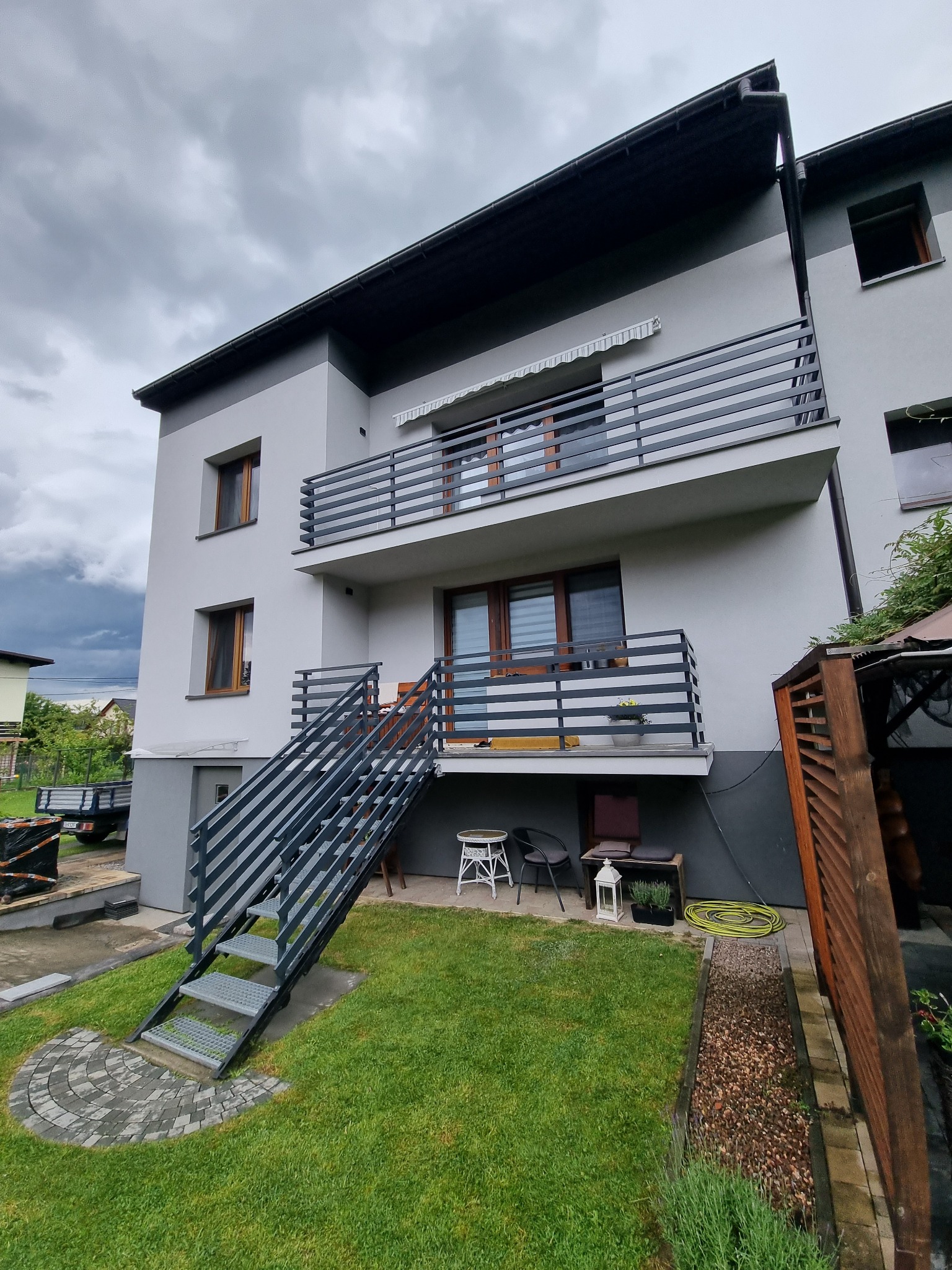 Nowoczesny, dwukondygnacyjny dom z szarą elewacją, balkonami z metalowymi balustradami i zewnętrznymi schodami z metalu prowadzącymi na wyższy poziom. Widoczny fragment ogrodu z trawnikiem...