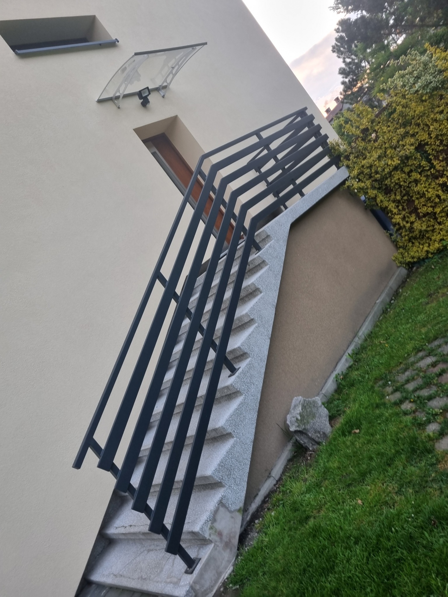 Zewnętrzne schody z szarymi stopniami i czarną metalową poręczą, prowadzące do drzwi wejściowych domu z kremową elewacją i szklanym daszkiem.