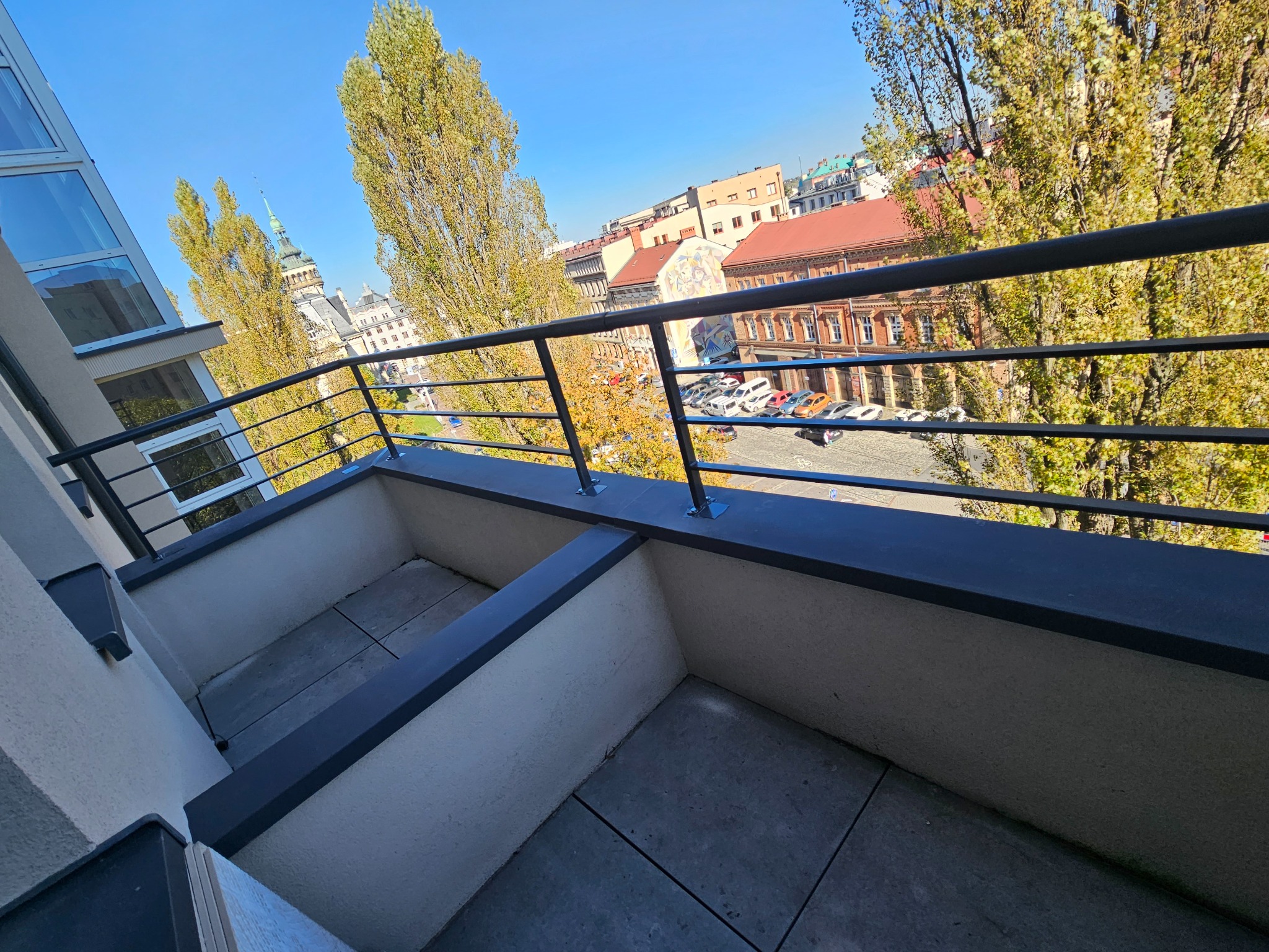 Balkon z czarną, metalową balustradą na tle jesiennego krajobrazu miejskiego z widokiem na zabytkowy budynek z wieżą.