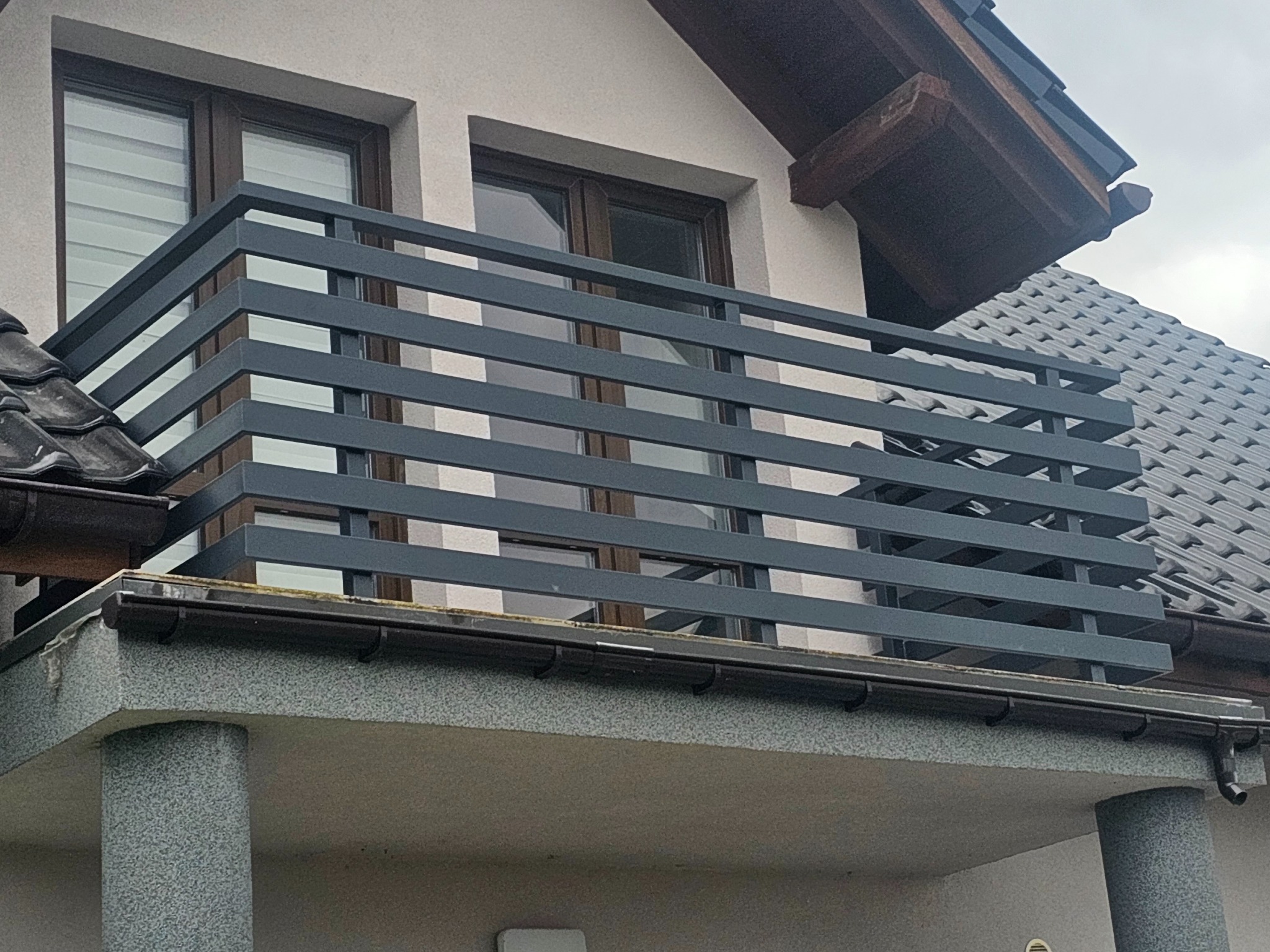 Balkon z nowoczesną, poziomą balustradą w kolorze grafitowym, widok z dołu, na tle szarej elewacji i dachu z dachówki ceramicznej.