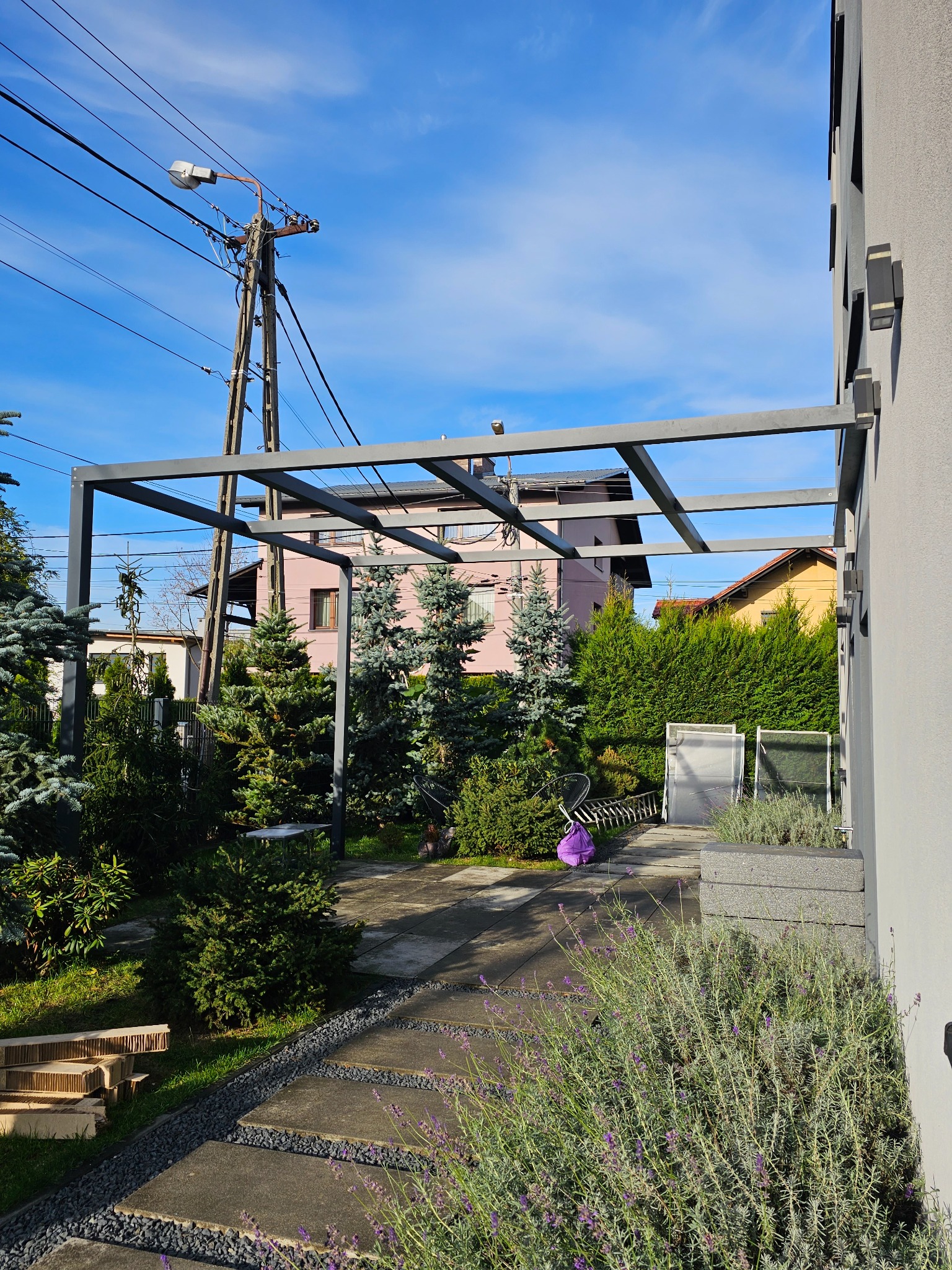 Szara metalowa konstrukcja pergoli w ogrodzie, widok na zadbany trawnik, ścieżkę z płyt i dom z różową elewacją w tle, błękitne niebo.