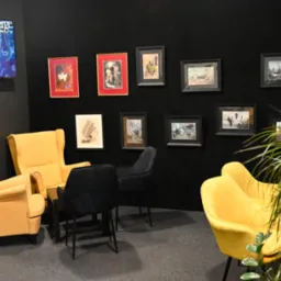 Żółte fotele klubowe i czarne krzesła na tle czarnej ściany z grafikami w ramkach, telewizor z logo Concierge w rogu, roślina doniczkowa w prawym dolnym rogu.