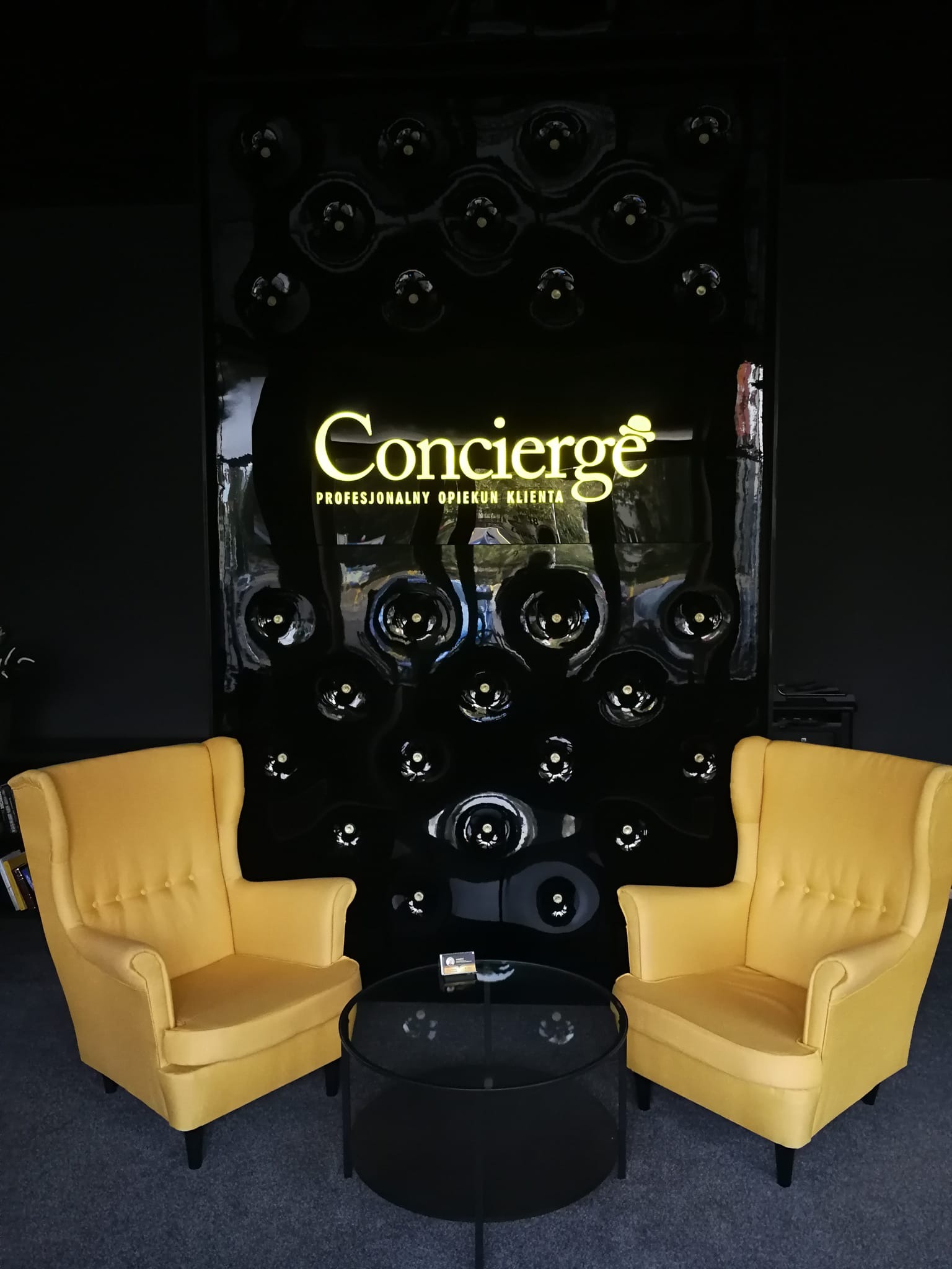 Żółte fotele klubowe i czarny stolik kawowy na tle ściany z logo 'Concierge' i dekoracyjnymi elementami w kształcie butelek, tworzące elegancką przestrzeń recepcyjną.