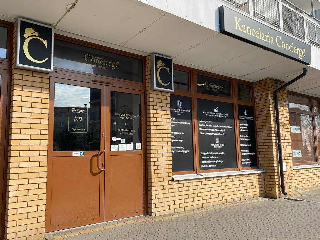 Fasada budynku z szyldem 'Kancelaria Concierge Prawo i Finanse', brązowe drzwi wejściowe z informacją o godzinach otwarcia i numerach telefonów, okna z wypisanymi usługami, ceglana elewacja.