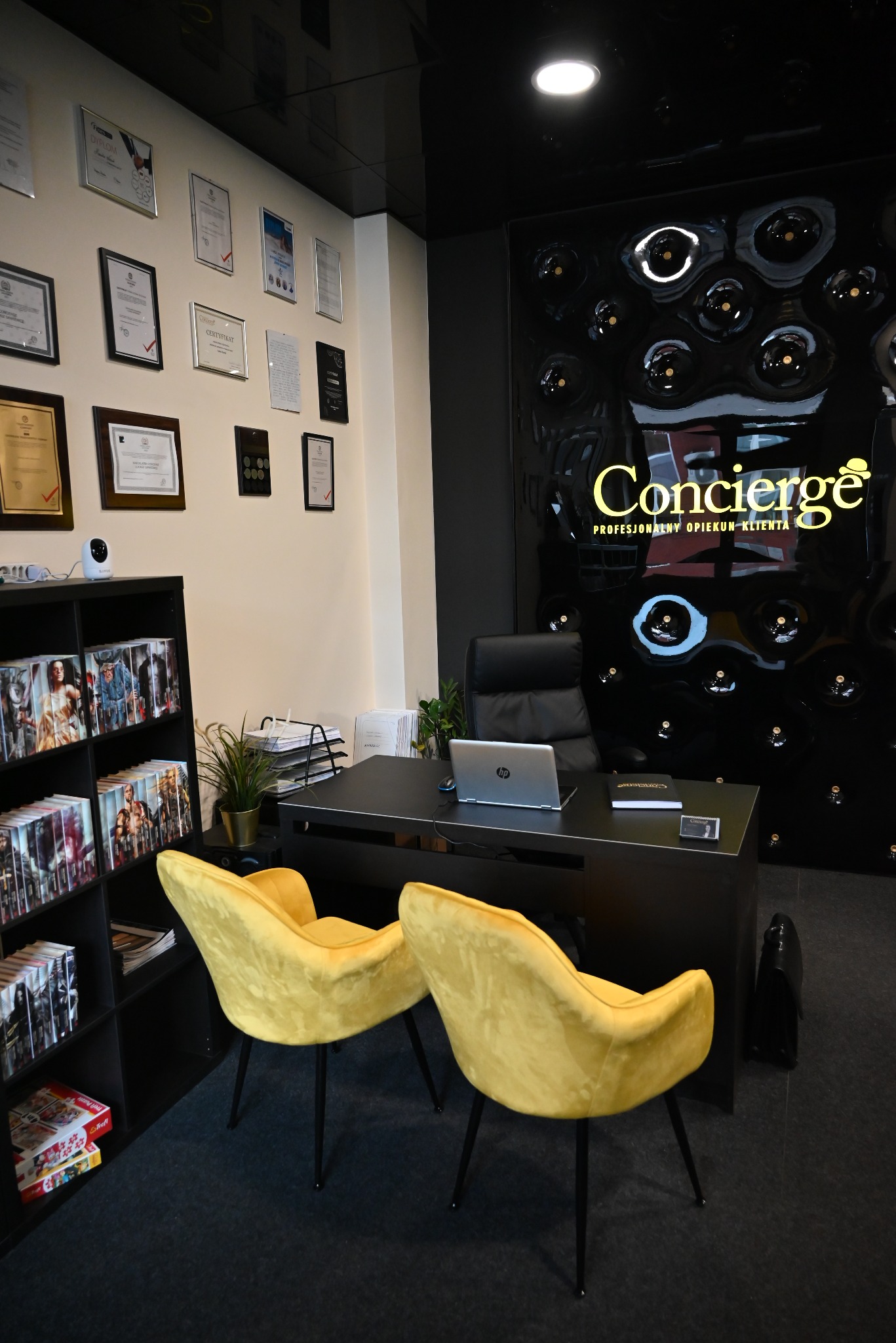 Nowoczesne biuro w Łomży z czarnym biurkiem, laptopem, dwoma żółtymi krzesłami i ścianą z certyfikatami oraz logo firmy 'Concierge'.
