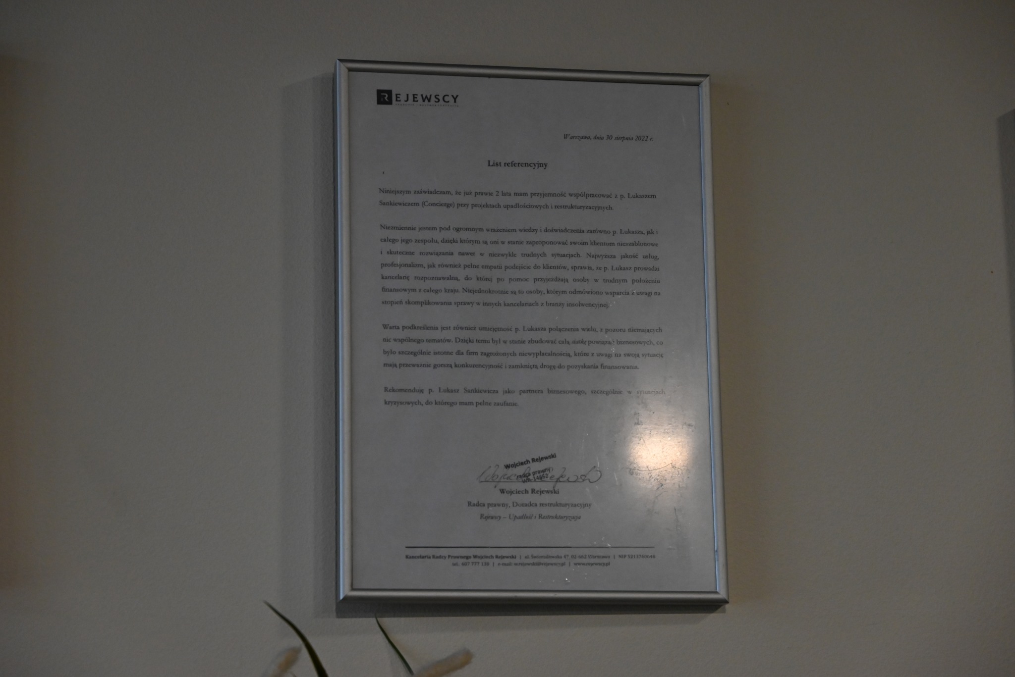 Oprawiony w ramkę list referencyjny kancelarii prawnej Rejewscy, datowany na 30 sierpnia 2022, z rekomendacją dla Łukasza Sankiewicza, zawierający podpis Wojciecha Rejewskiego, radcy prawnego...