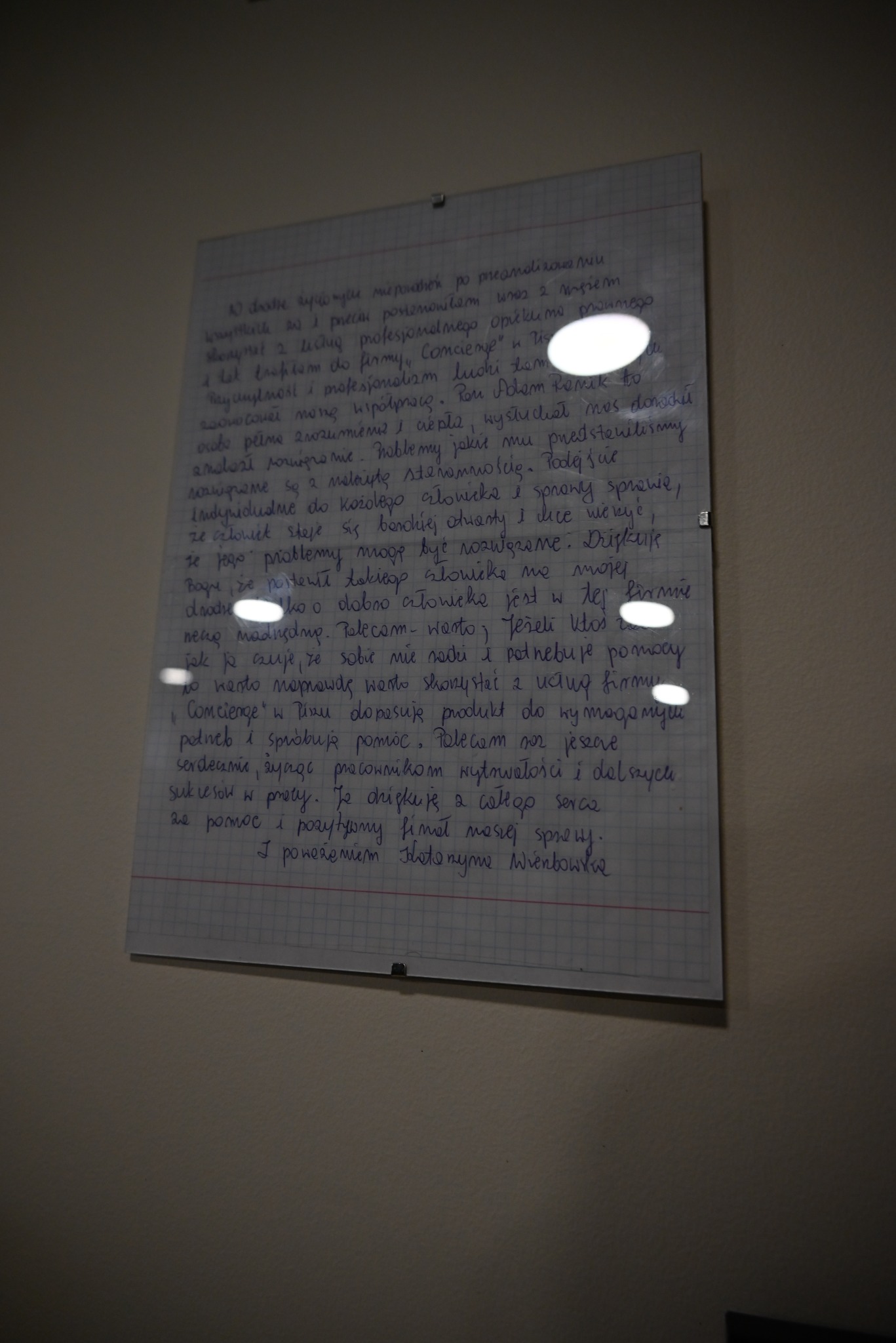 Ręcznie napisane podziękowanie na kartce w kratkę, przytwierdzone do ściany, z tekstem polecającym firmę Concierge i jej profesjonalizm, z widocznymi refleksami światła.