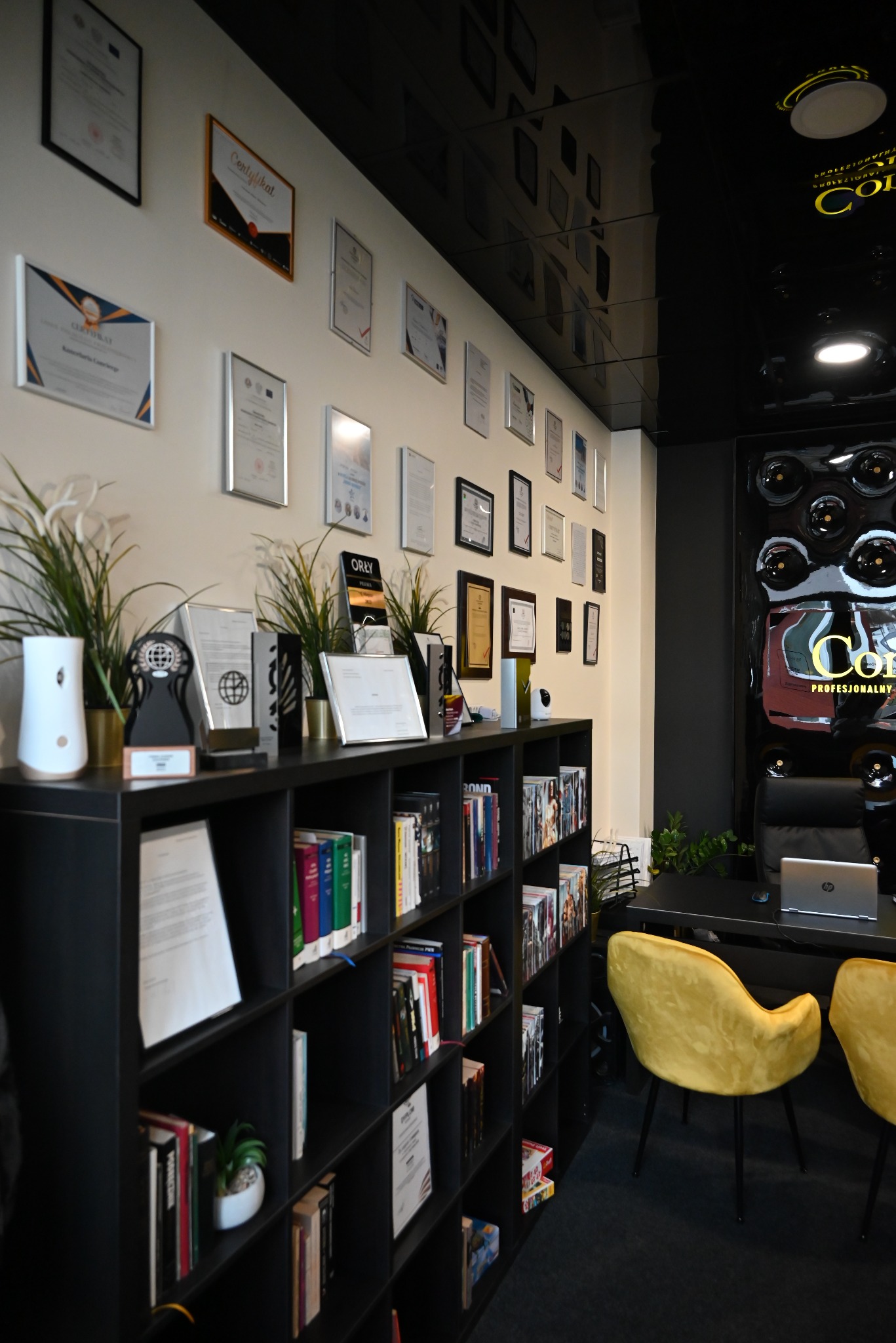 Wnętrze biura z czarnymi regałami wypełnionymi książkami i dokumentami, na ścianie liczne certyfikaty i nagrody, biurko z laptopem i złote krzesła.