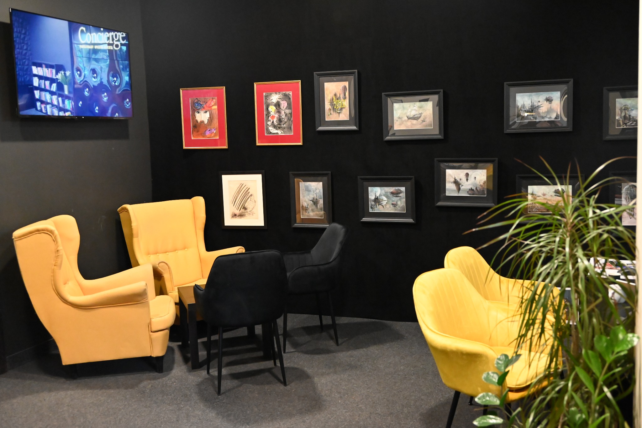 Żółte fotele klubowe i czarne krzesła na tle czarnej ściany z grafikami w ramkach, telewizor z logo Concierge w rogu, roślina doniczkowa w prawym dolnym rogu.