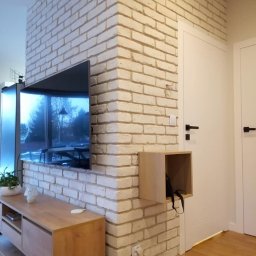 Serra's team Zakład Budowlany Rafał Serafin - Telewizor wkomponowany w ścianę z białej cegły, drewniana szafka RTV, drewniana podłoga, białe drzwi z czarnymi klamkami i mała półka w ścianie.