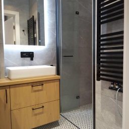 Serra's team Zakład Budowlany Rafał Serafin - Nowoczesna łazienka z drewnianą szafką pod umywalką, kabiną prysznicową ze szklanymi drzwiami i czarnym grzejnikiem drabinkowym na tle szarych płytek imitujących marmur i wzorzystej podłogi.