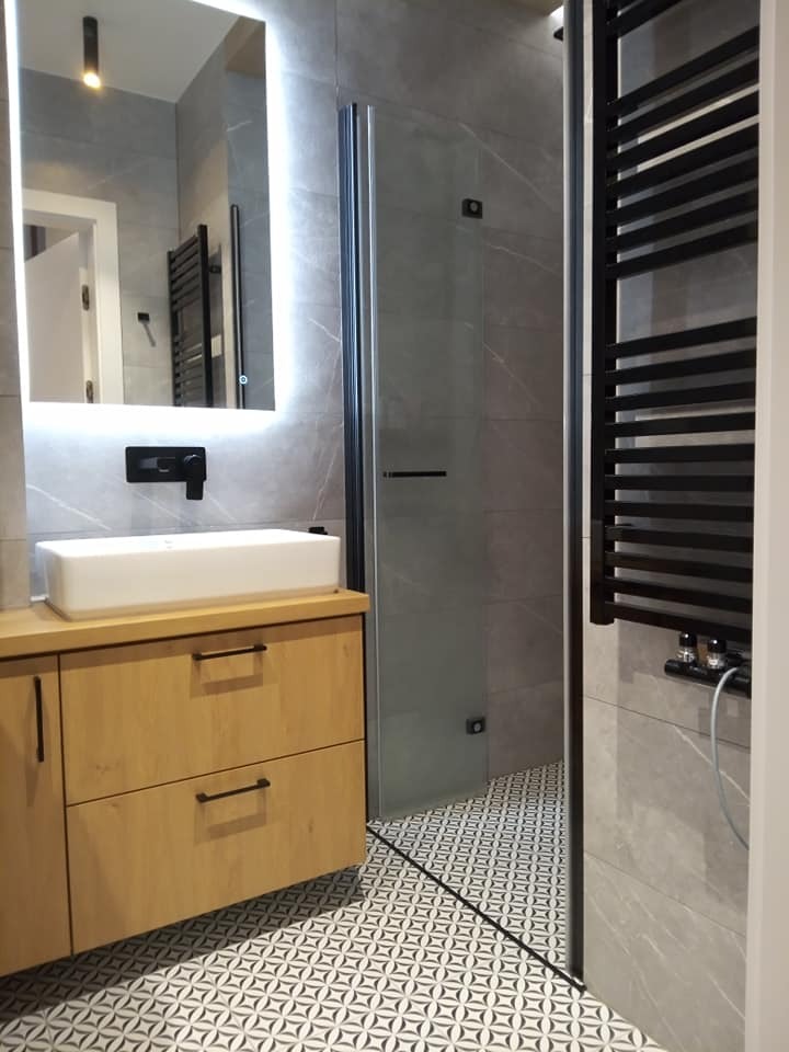 Nowoczesna łazienka z drewnianą szafką pod umywalką, kabiną prysznicową ze szklanymi drzwiami i czarnym grzejnikiem drabinkowym na tle szarych płytek imitujących marmur i wzorzystej podłogi.