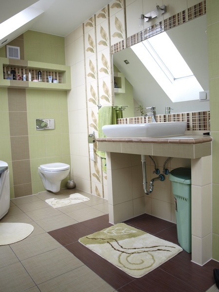 Jasna łazienka na poddaszu z umywalką nablatową, toaletą podwieszaną i zielonymi akcentami w postaci płytek z motywem liści, ręcznika i kosza na śmieci.