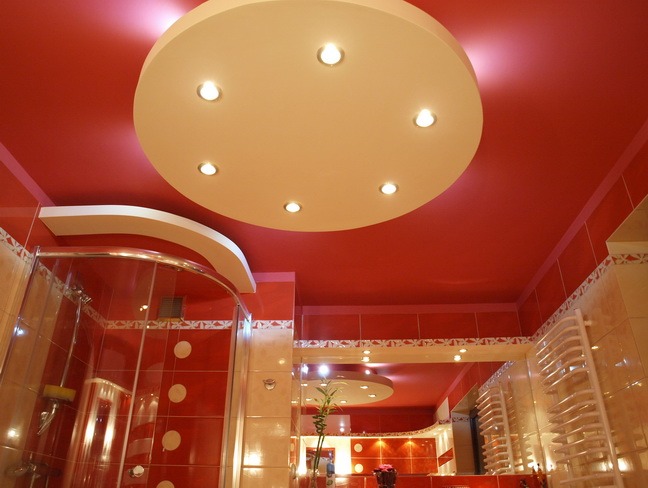 Czerwona łazienka z okrągłym, podwieszanym sufitem z sześcioma punktami świetlnymi, widoczna kabina prysznicowa z okrągłymi dekoracjami na kafelkach i grzejnik drabinkowy.